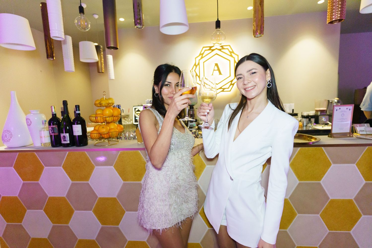 Two women in elegant attire toasting with champagne glasses at a stylish bar with honeycomb tile decor.