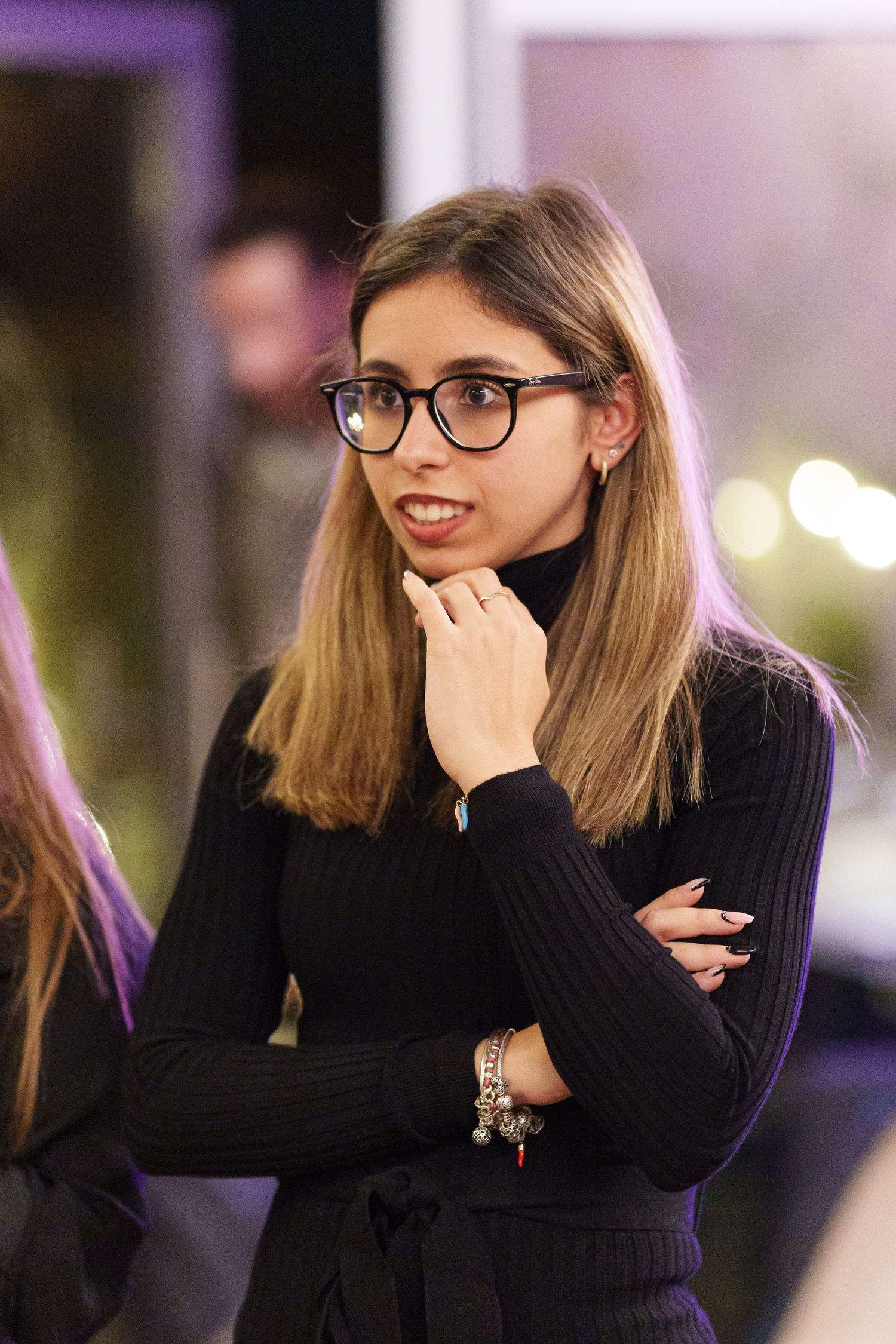 Young woman with glasses and long hair wearing black turtleneck, posing thoughtfully at an evening event.