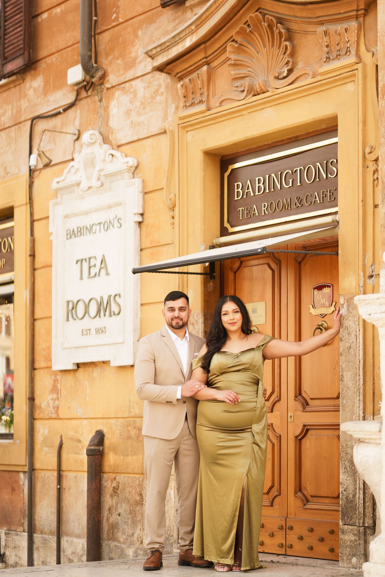 Pregnant couple posing in elegant attire outside Babington's Tea Rooms in Rome, Italy.