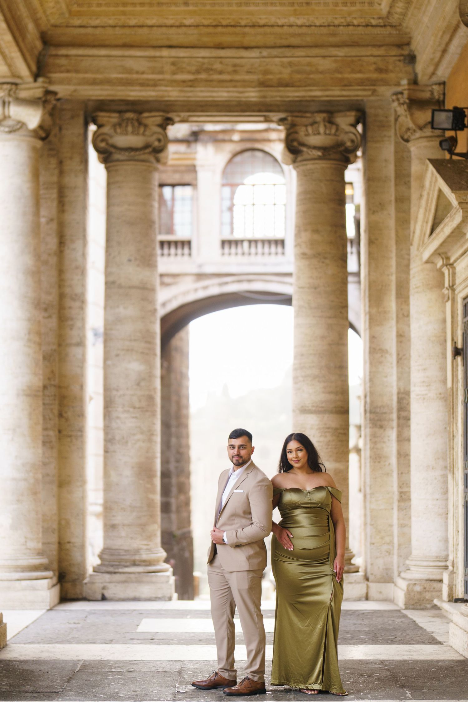 Couple posing elegantly between grand stone columns, man in beige suit and woman in olive green gown, Rome architecture.
