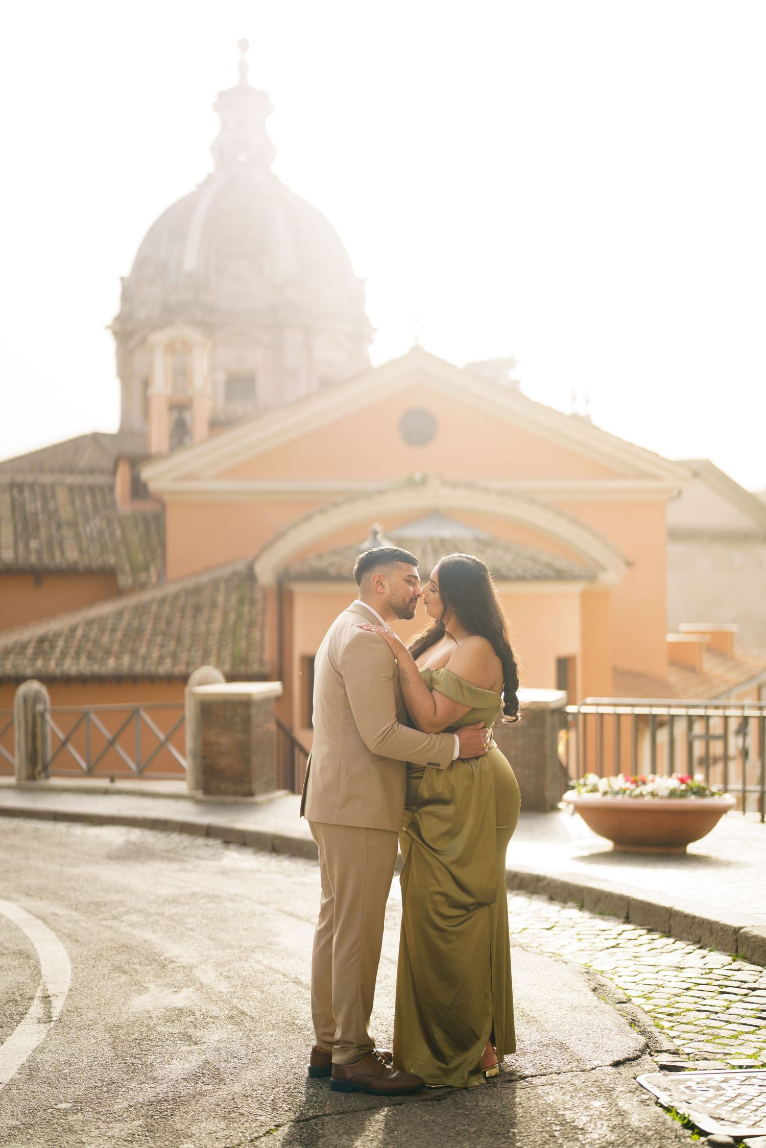 Couple embracing near a Roman church dome at golden hour, man in tan suit, woman in olive green dress.