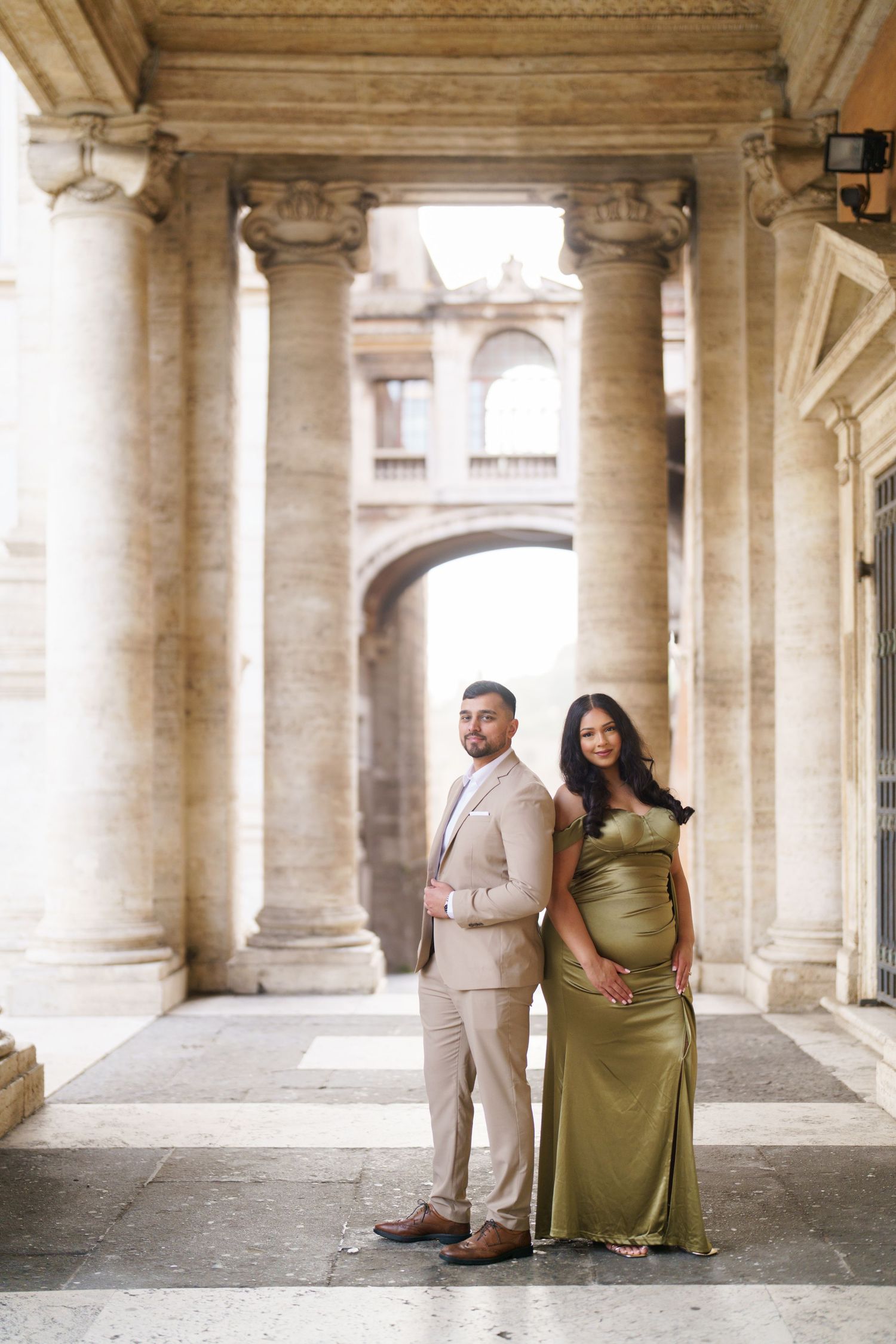 Elegant couple posing among grand stone columns, woman in olive satin gown and man in beige suit, classical architecture backdrop.