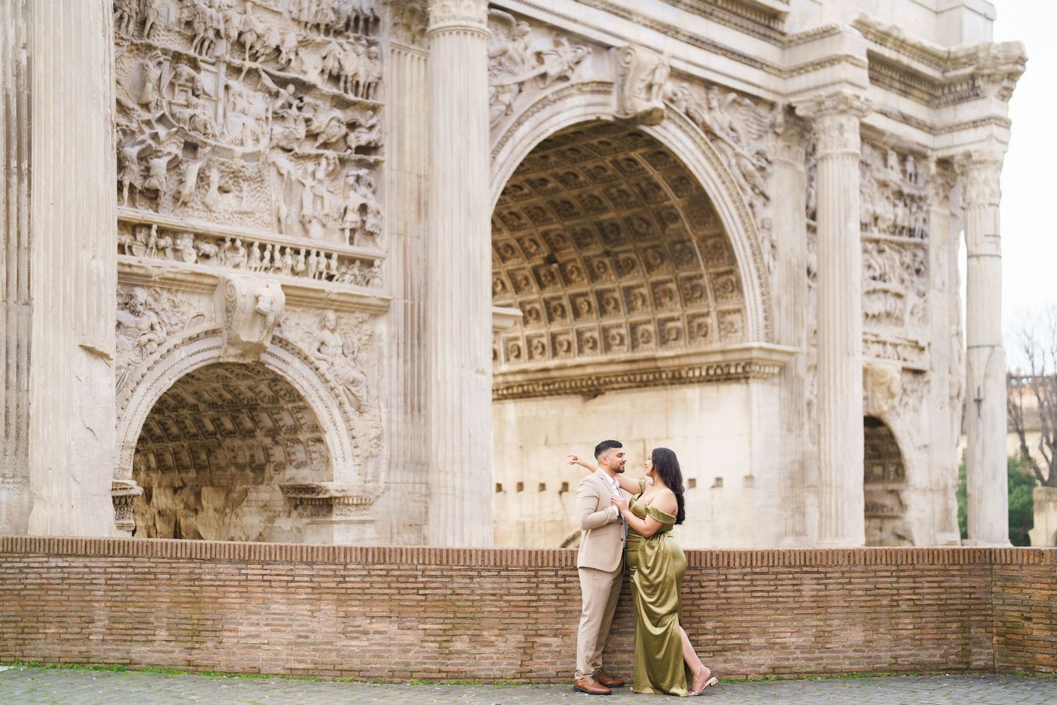 Couple embracing in front of the Arch of Constantine in Rome, woman in gold dress, man in beige suit.