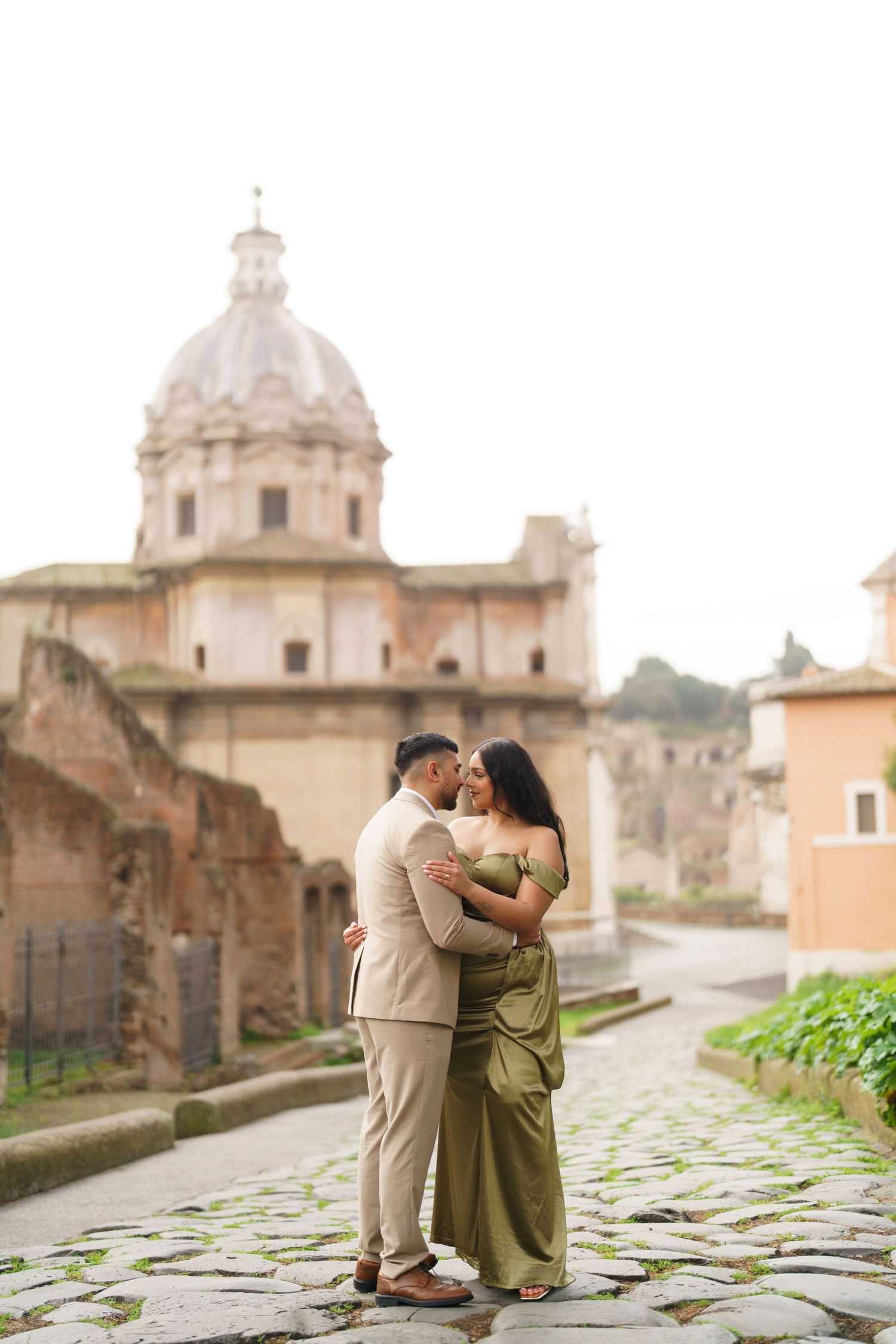 Couple embracing and kissing on cobblestone path with ancient Roman church dome backdrop in Rome, Italy.