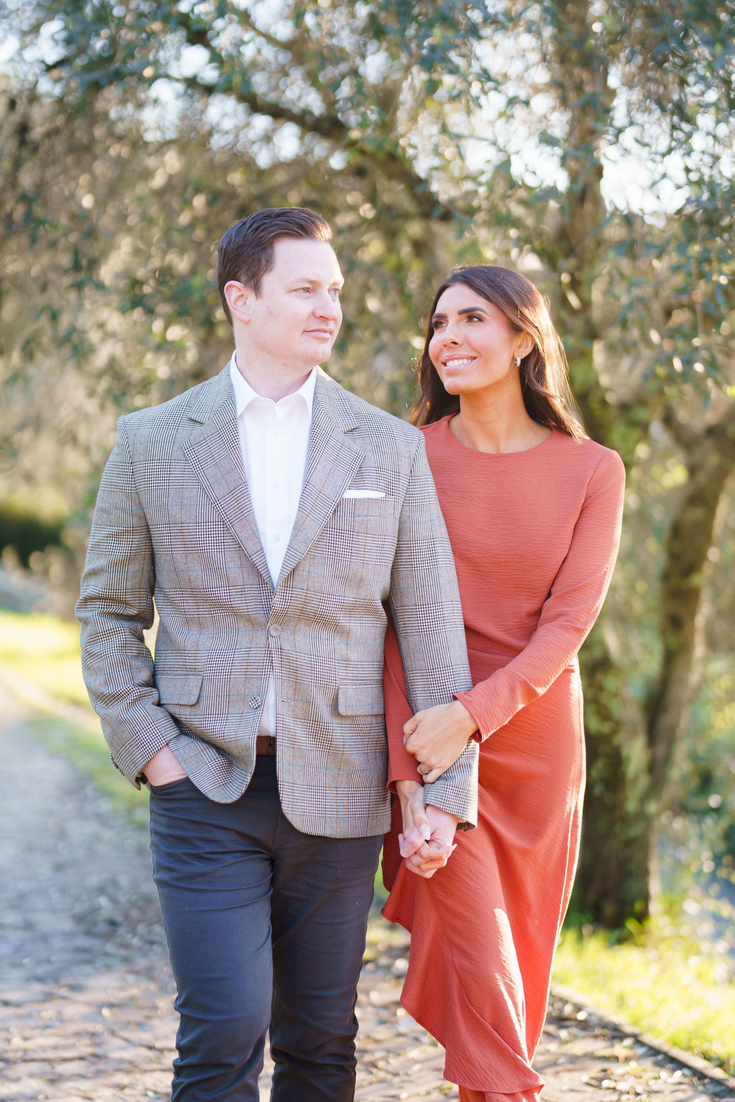 Couple holding hands walking outdoors, man in gray blazer, woman in rust dress, trees in background.