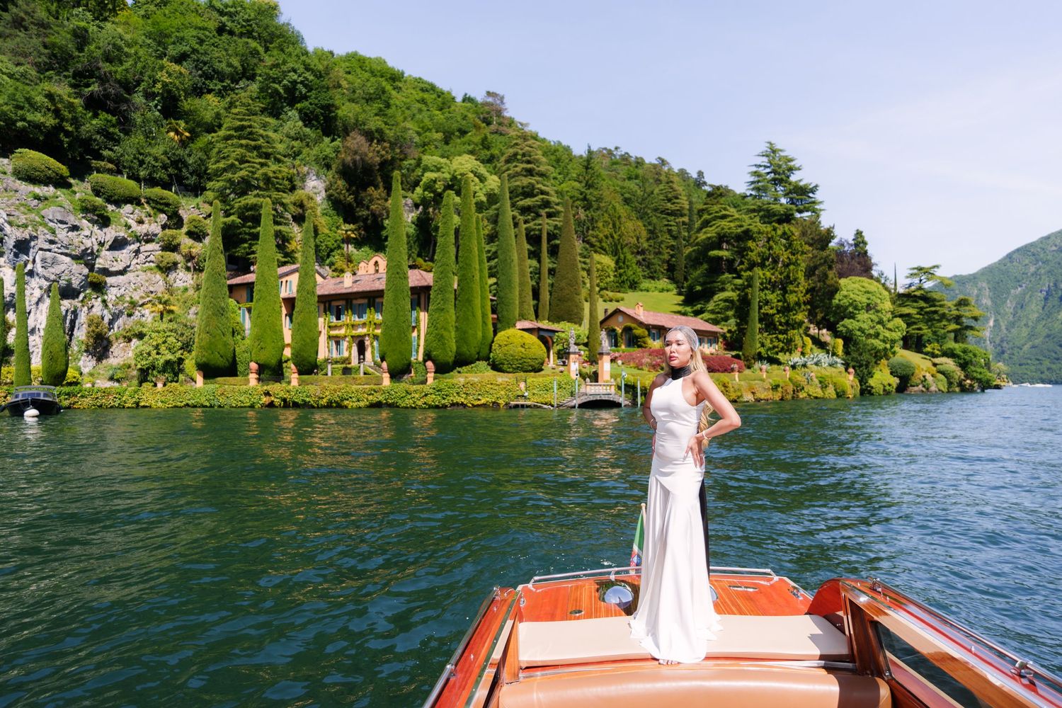 Bride in white gown poses on wooden boat bow with Lake Como villa and cypress trees in background.