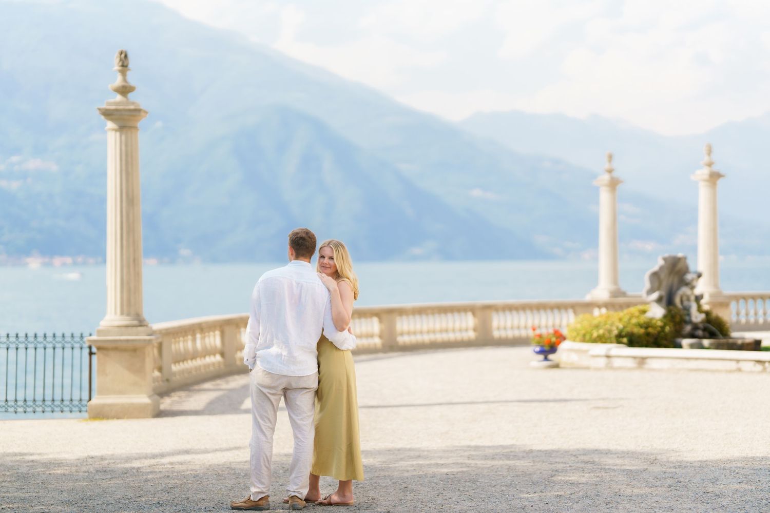 Couple embracing on elegant lakeside terrace with mountain views and classical columns at Lake Como, Italy.