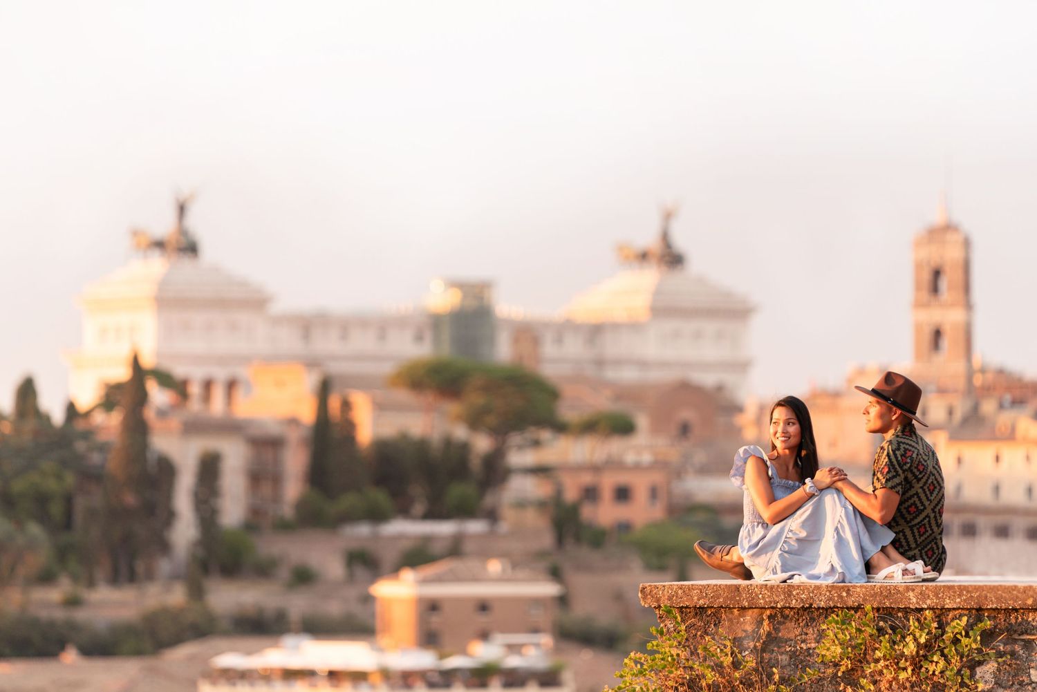 Romantic couple sitting on a stone wall overlooking Rome's historic skyline at golden hour sunset.