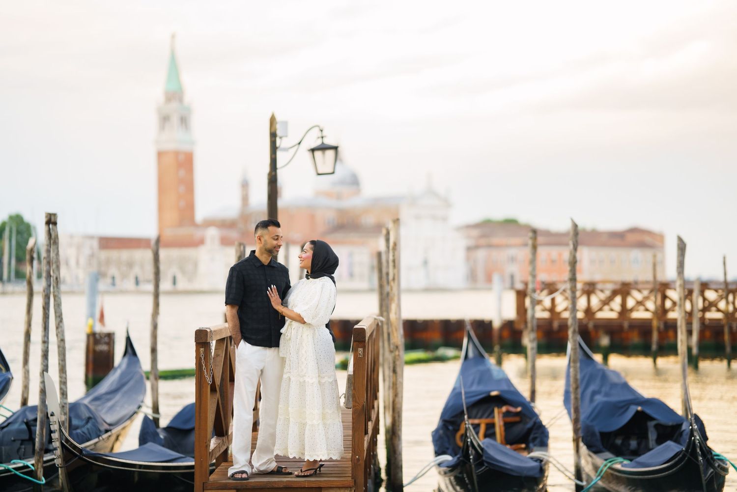 Couple embracing on a wooden dock in Venice, Italy, with gondolas and San Giorgio Maggiore island in the background.