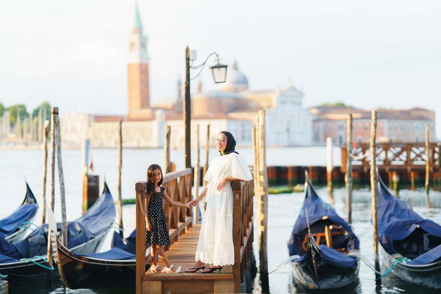 Two women hold hands on a Venice gondola dock with San Giorgio Maggiore church in the background.