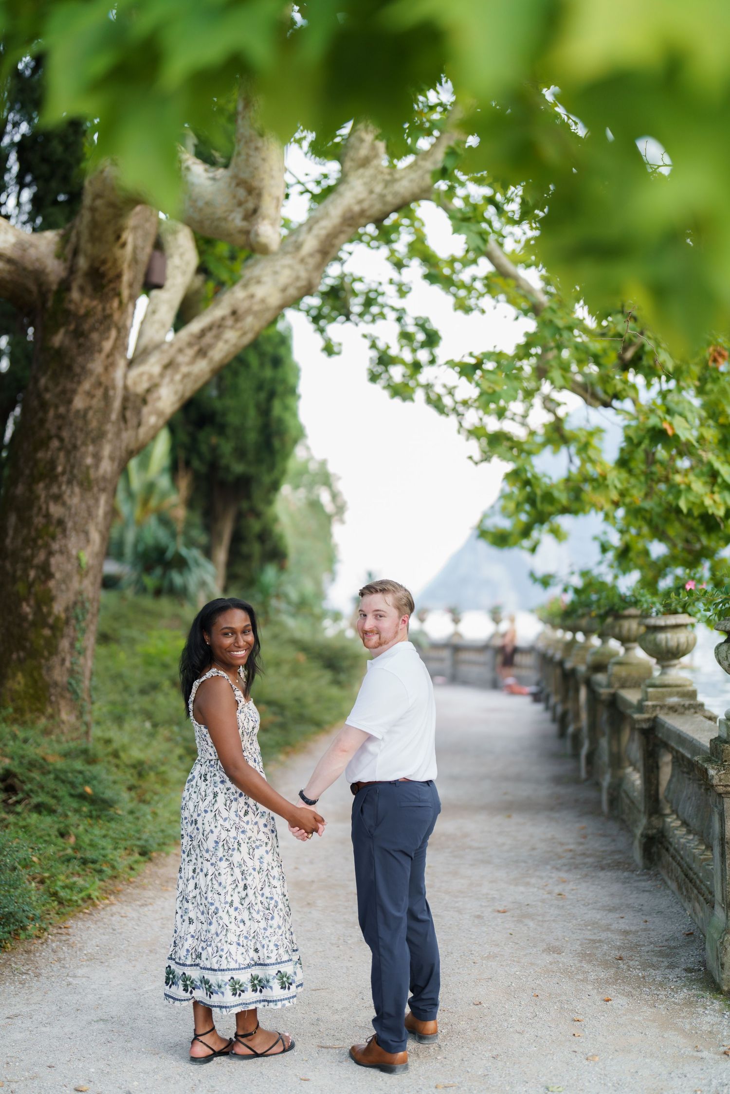 Interracial couple holding hands on a scenic garden path lined with trees and stone balustrade.