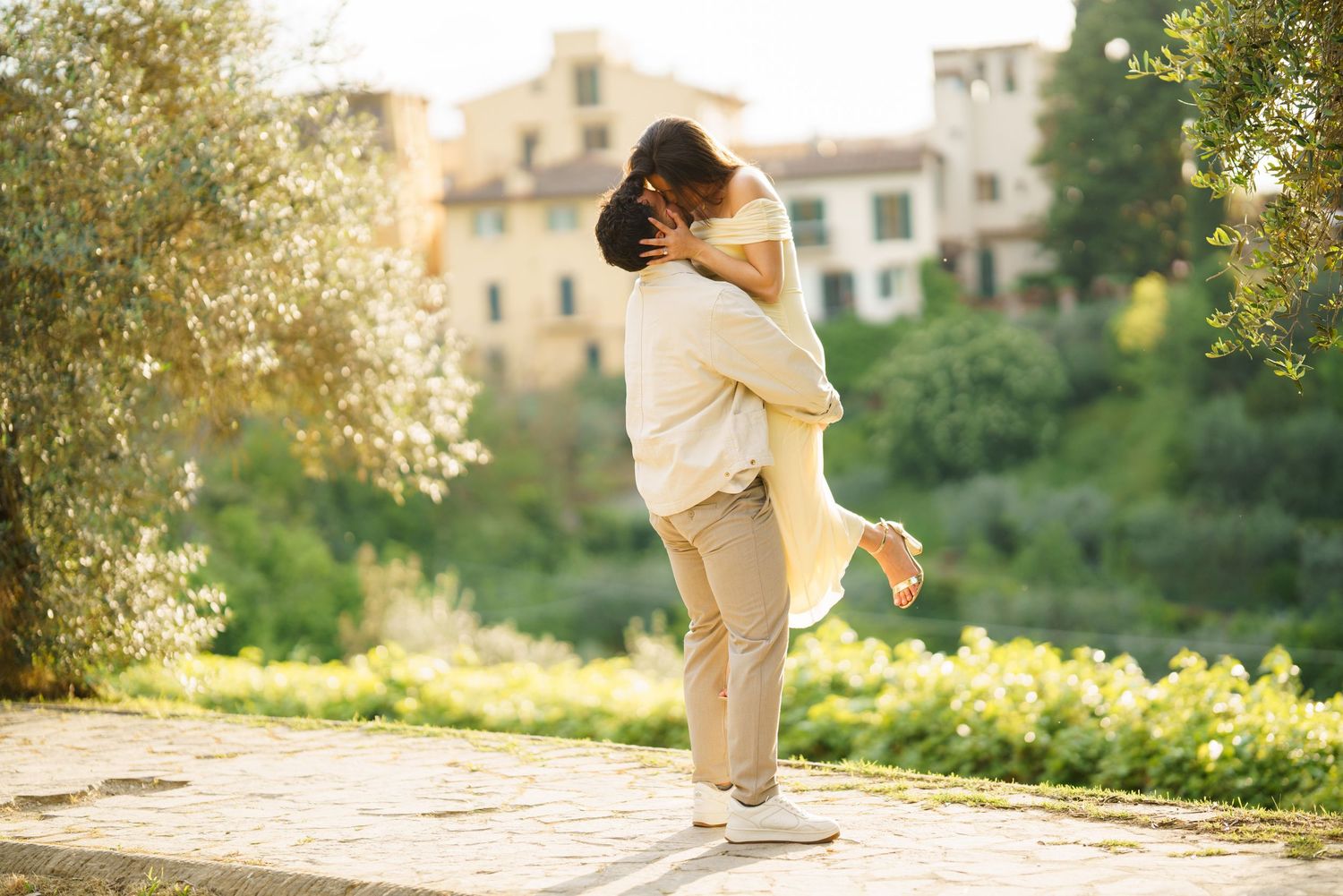 Couple embracing romantically in a sunlit Italian garden, man lifting woman in a joyful hug outdoors.