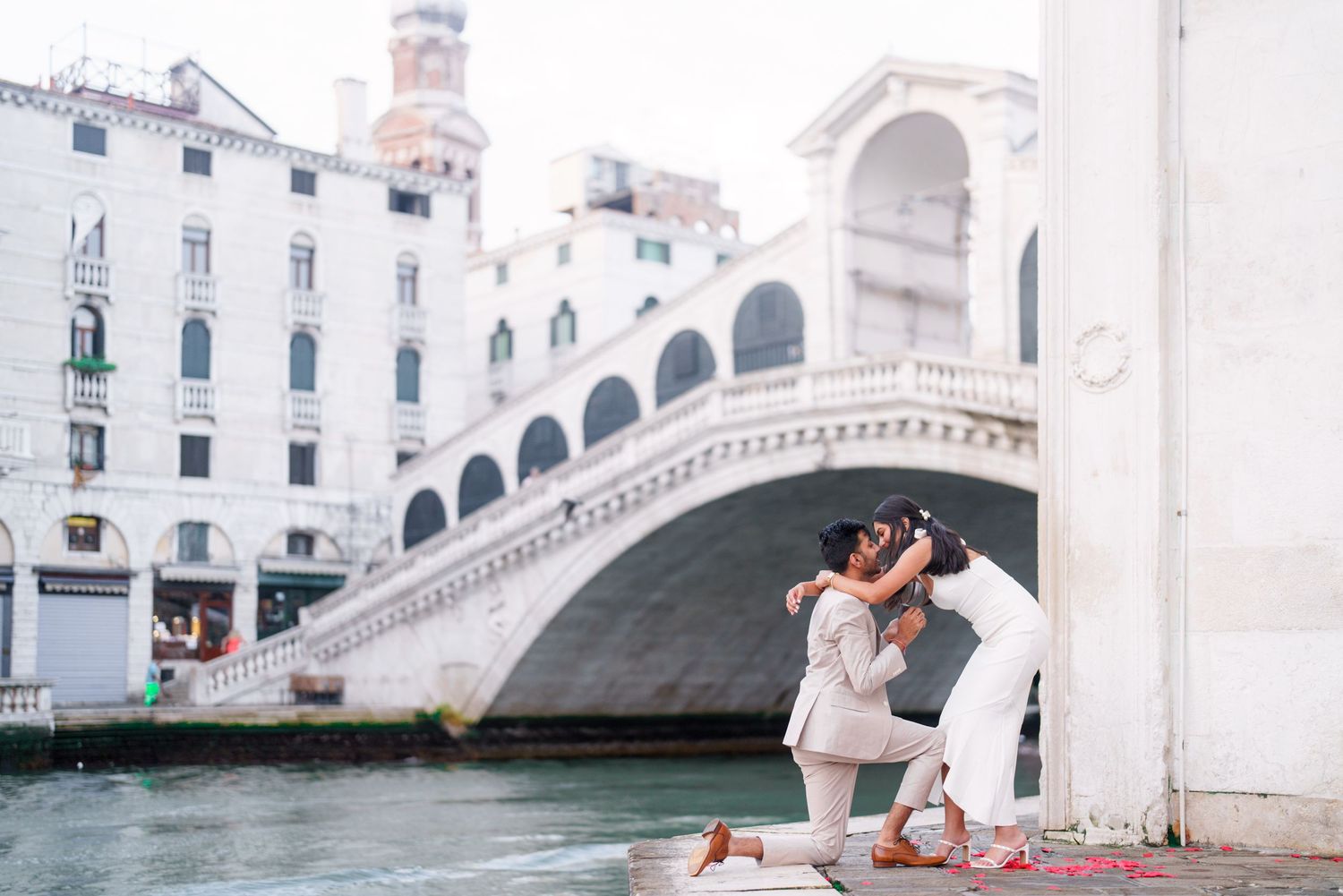 A romantic couple embraces on a bridge in Venice, Italy with classic architecture and canals in the background.