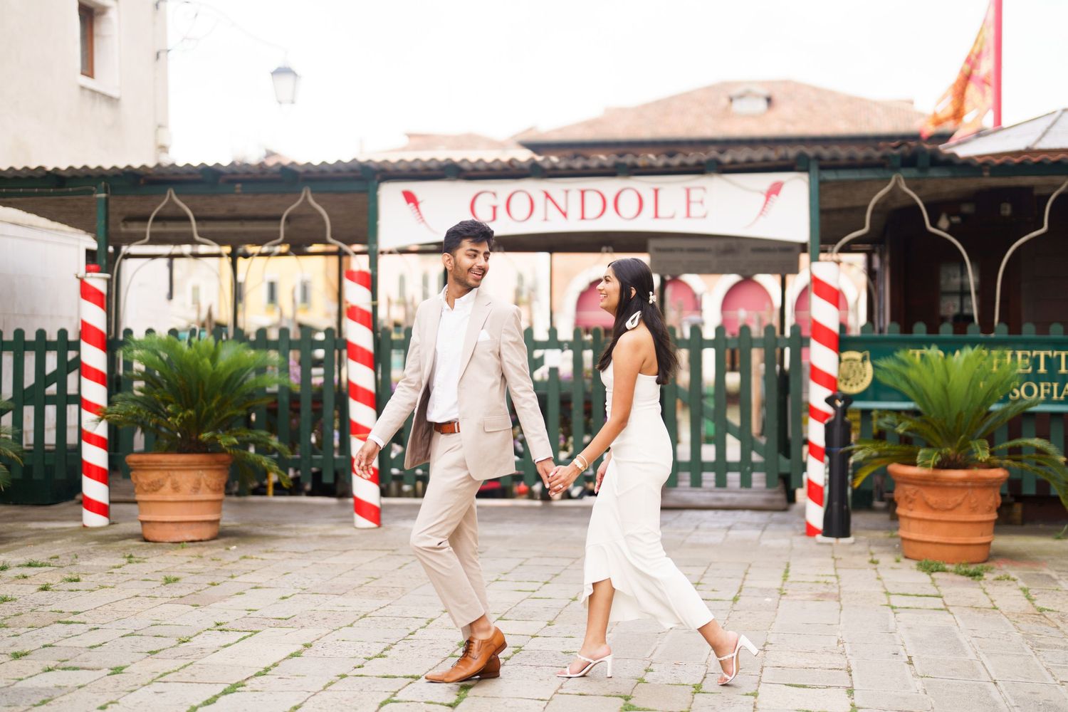 A person in a white dress walks along a rustic European street lined with red and white striped poles.