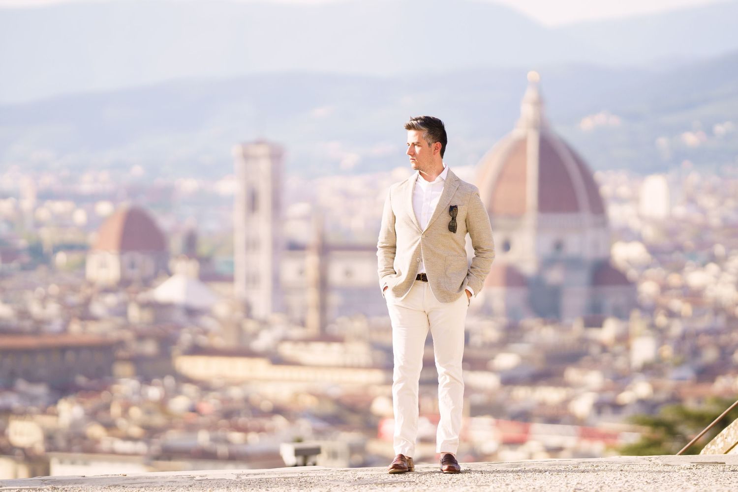 A person in white clothing poses against the scenic backdrop of Florence, Italy with the Duomo cathedral visible in the distance.