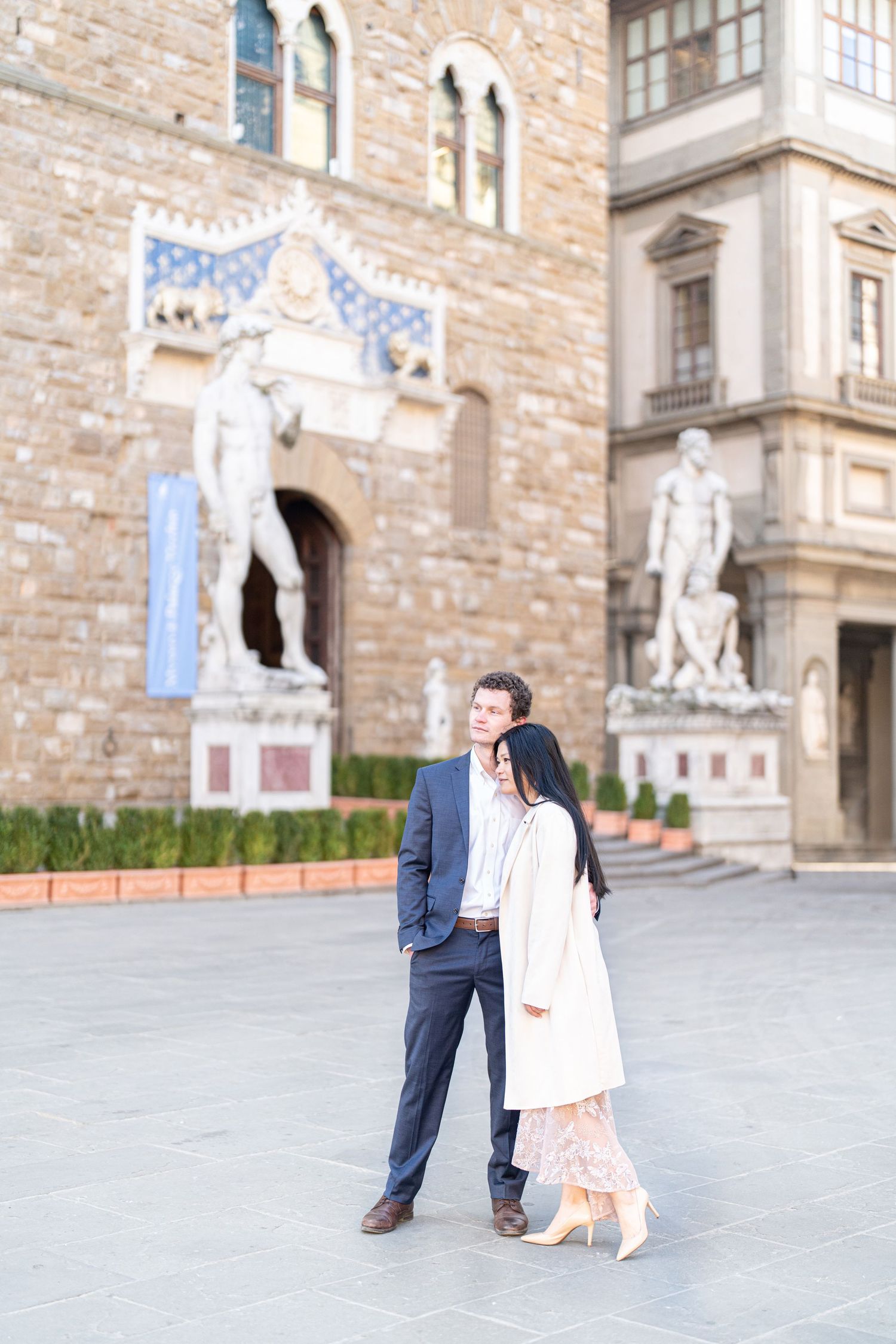 A couple stands together in front of historic Palazzo Vecchio in Florence, Italy with Renaissance statues in background.