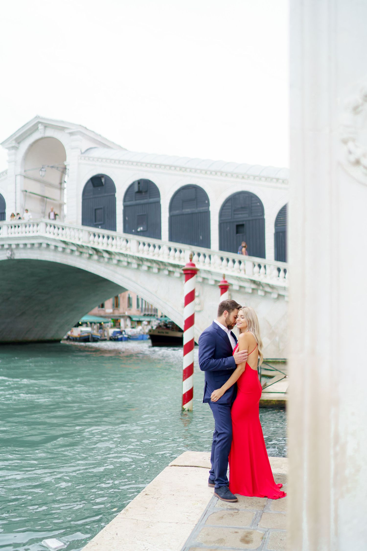 A romantic couple embraces near the iconic Rialto Bridge in Venice, Italy with the Grand Canal's turquoise waters behind them.
