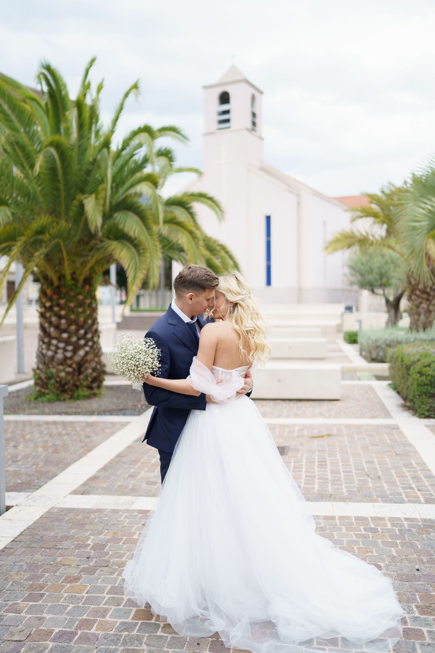 A romantic wedding couple shares a kiss in a Mediterranean courtyard with palm trees and brick pavers.