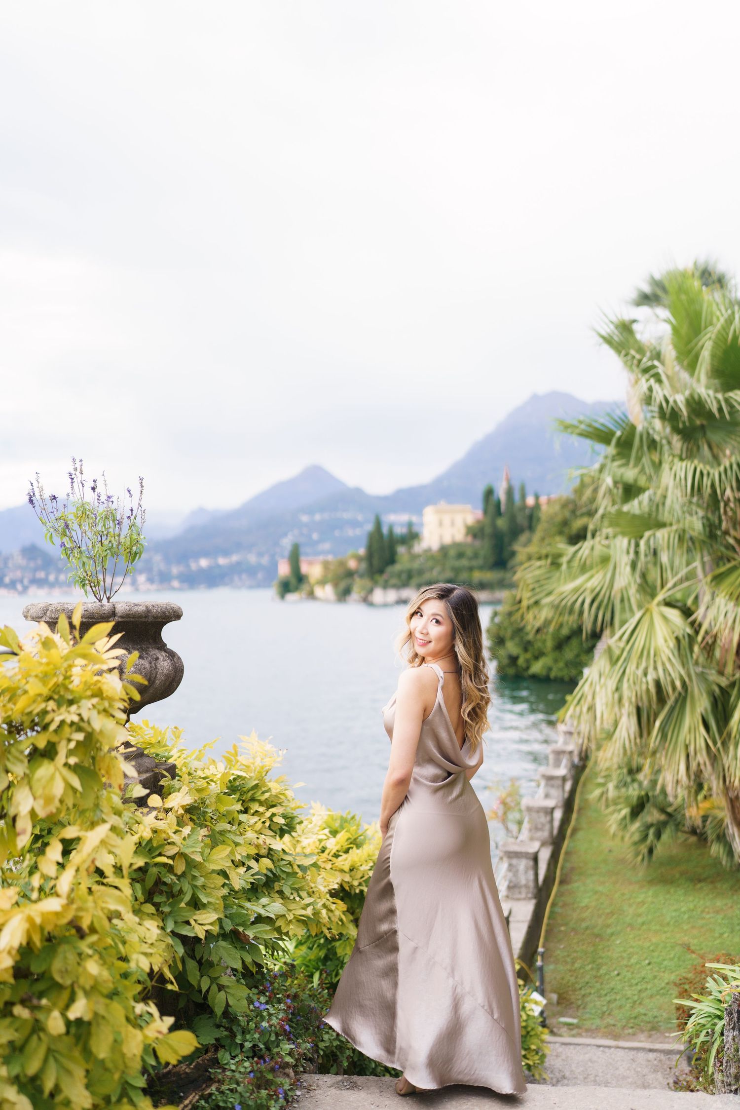 A woman in a beige dress poses near a lake with mountains and palm trees in a scenic Italian landscape setting.