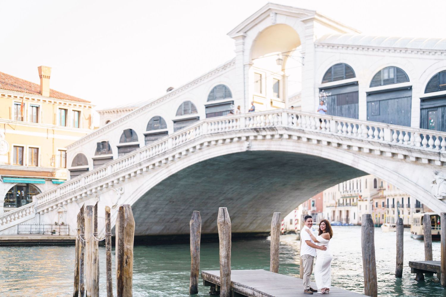 The iconic Rialto Bridge spans the Grand Canal in Venice, with its white stone arches reflecting in the blue waters below.