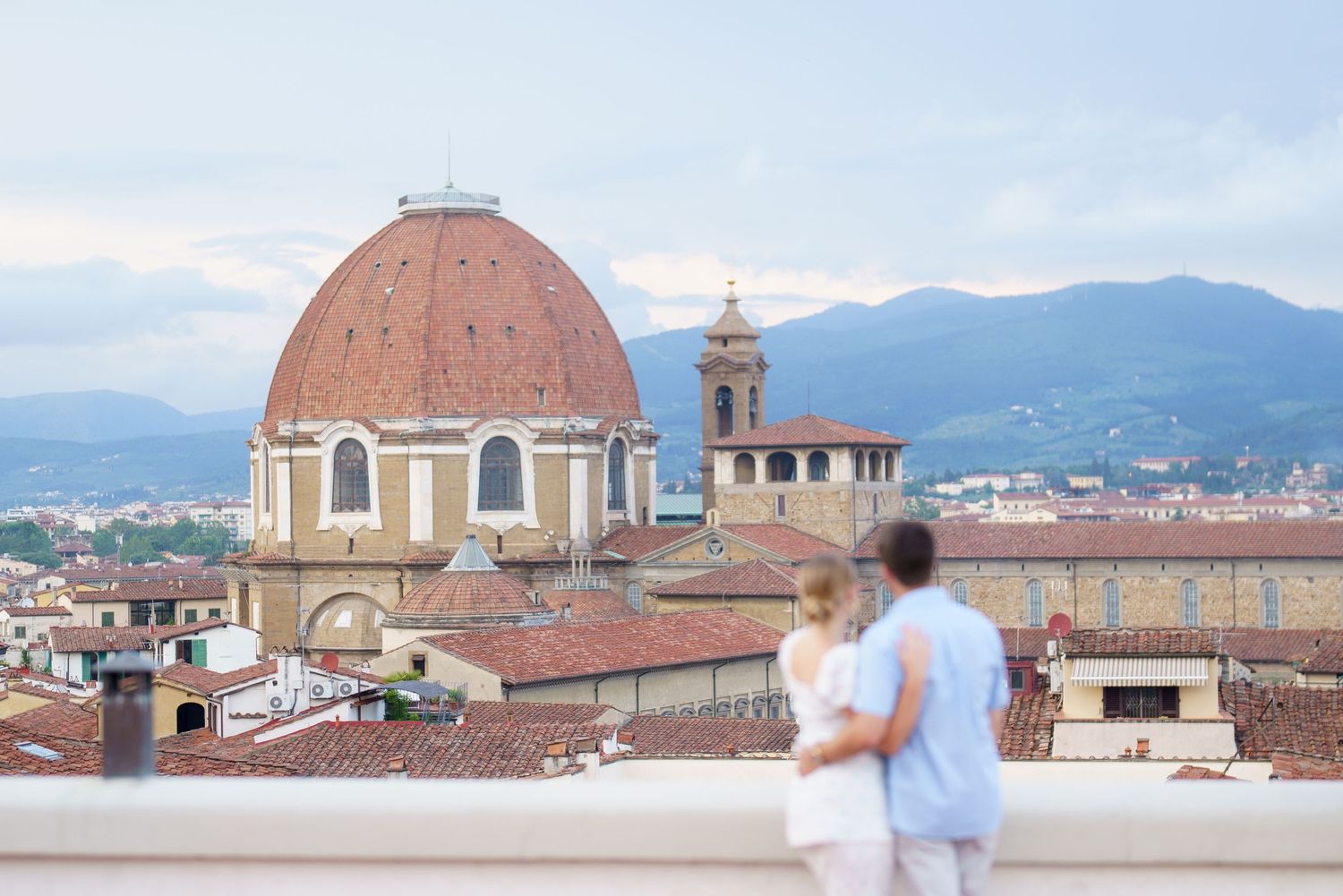 Couple admiring Florence cityscape with Medici Chapel dome and mountains in background.