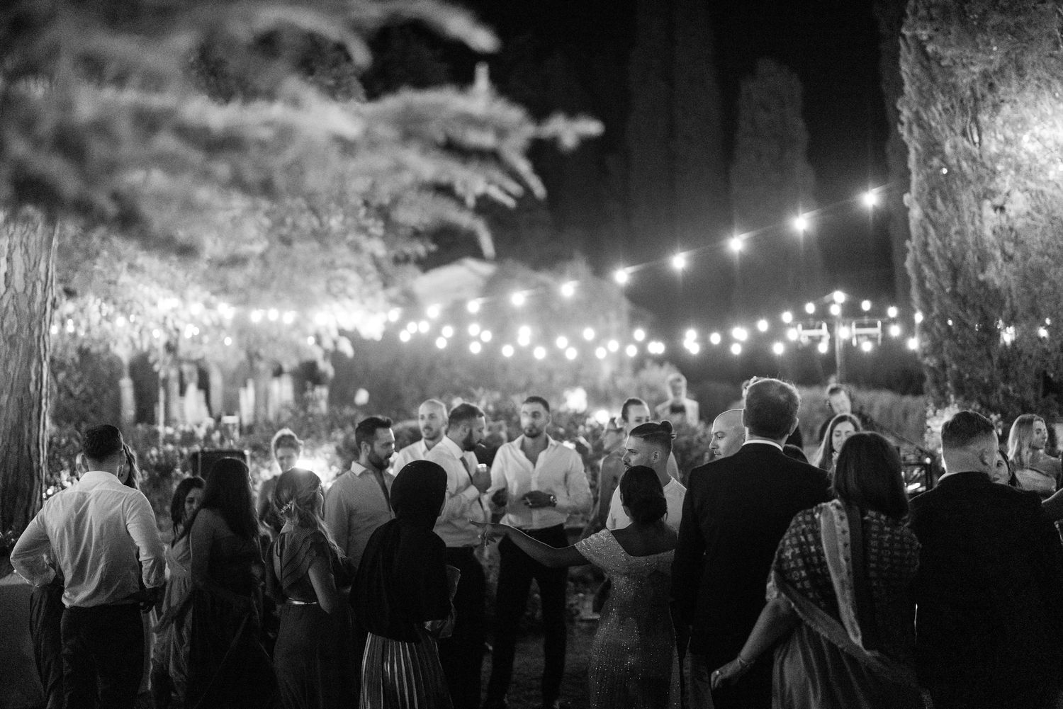 String lights illuminate a crowd of wedding guests dancing at an outdoor nighttime celebration.
