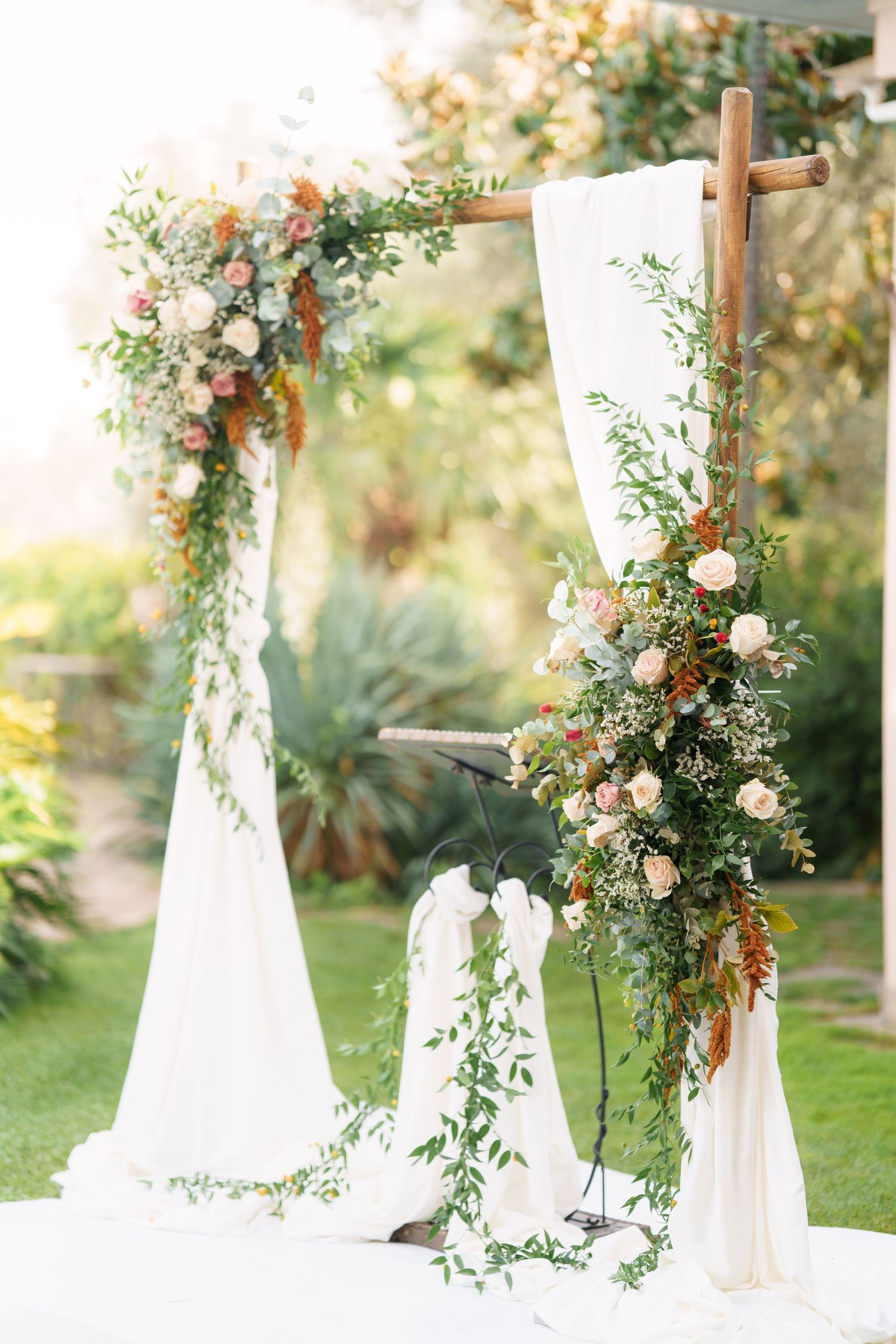 A wooden cross arch draped with white fabric and adorned with cascading flowers and greenery.