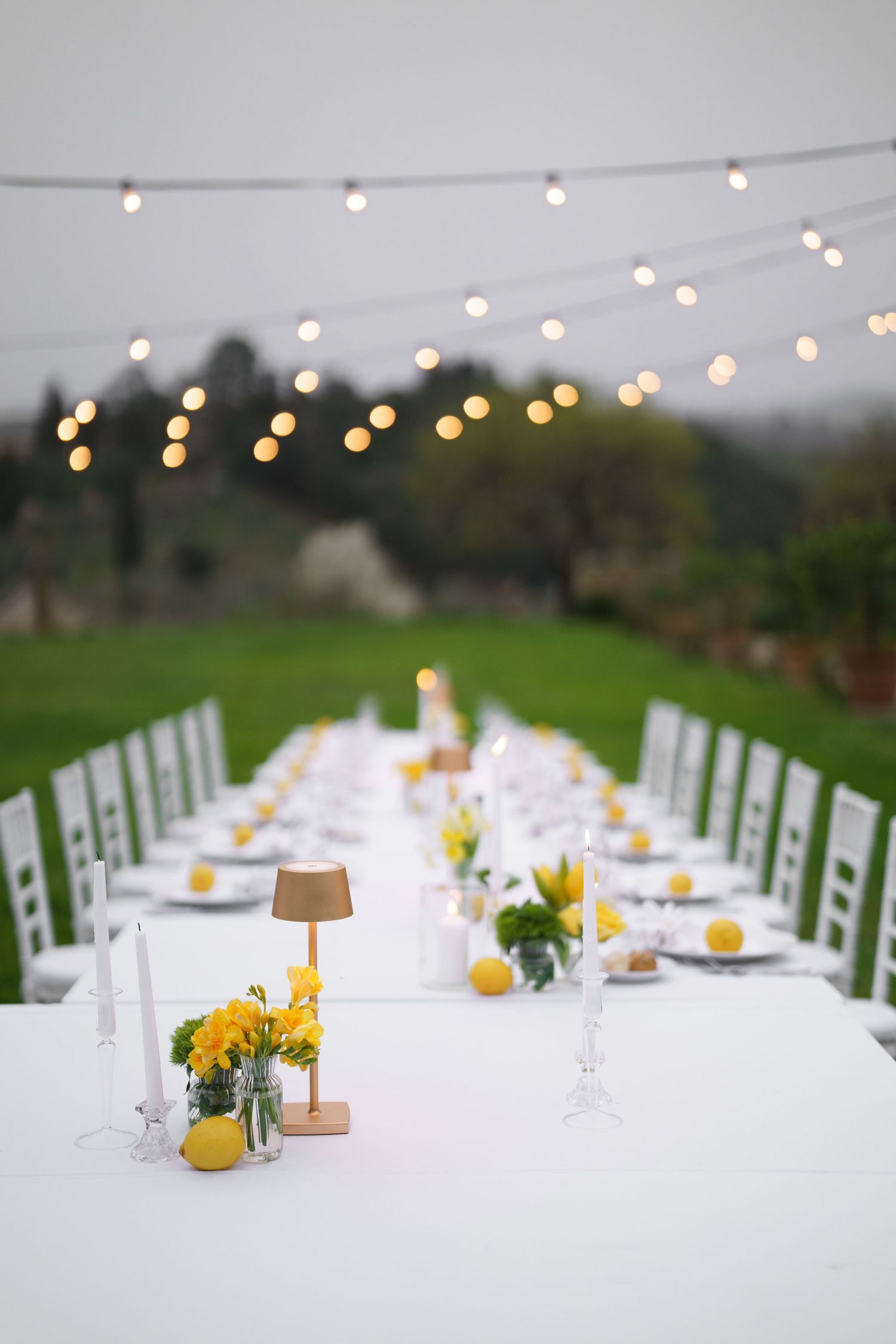 Long white dining table under string lights with yellow floral centerpieces and white chairs.