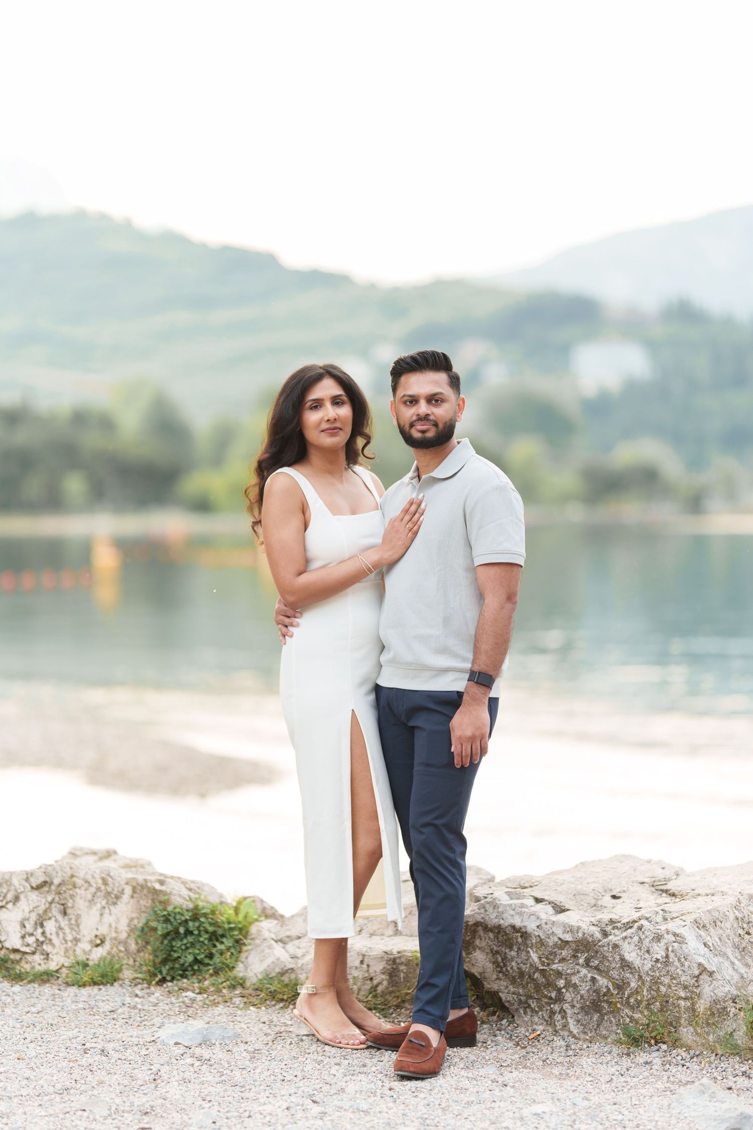 A couple poses together on a rocky shore with a serene lake and mountains in the background.