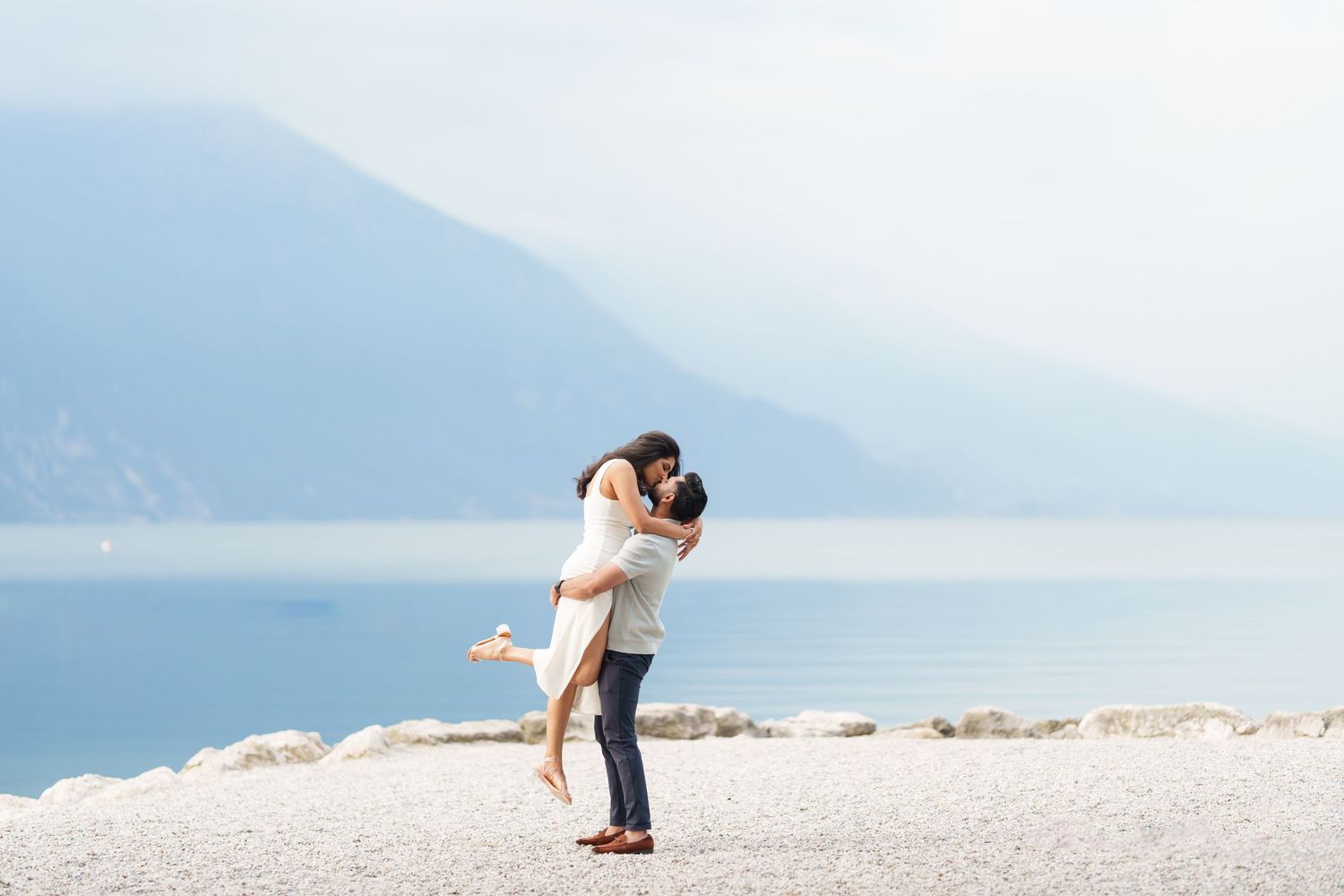 Two people share a romantic moment on a beach with mountains and ocean in the background.