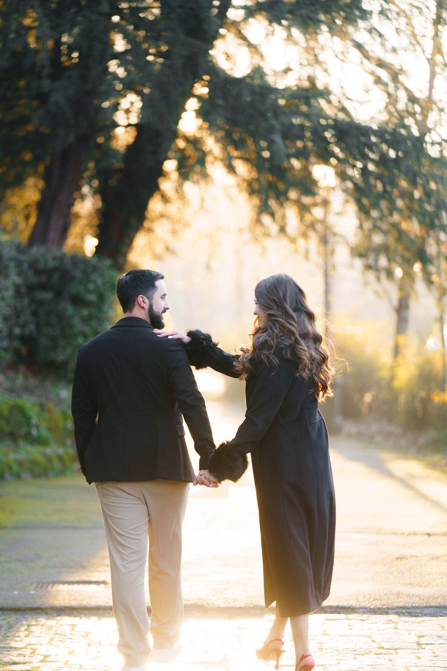 A couple walks hand in hand on a sunlit path through trees during golden hour creating a romantic silhouette.