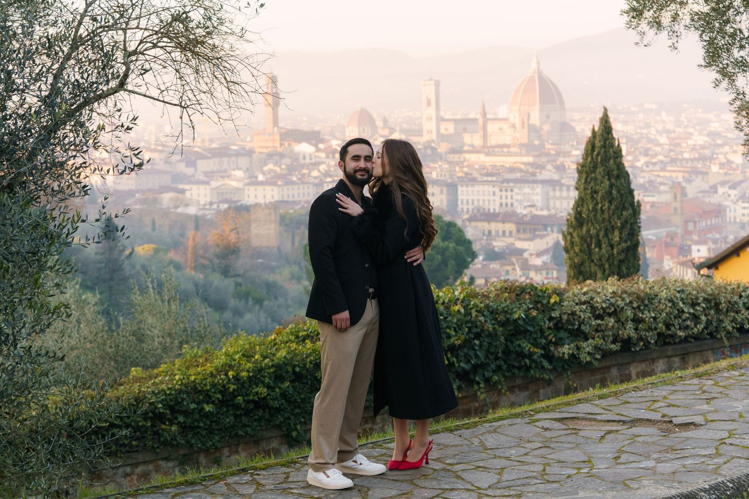 A couple shares a romantic moment at a scenic overlook with Florence's iconic Duomo cathedral visible in the misty background.