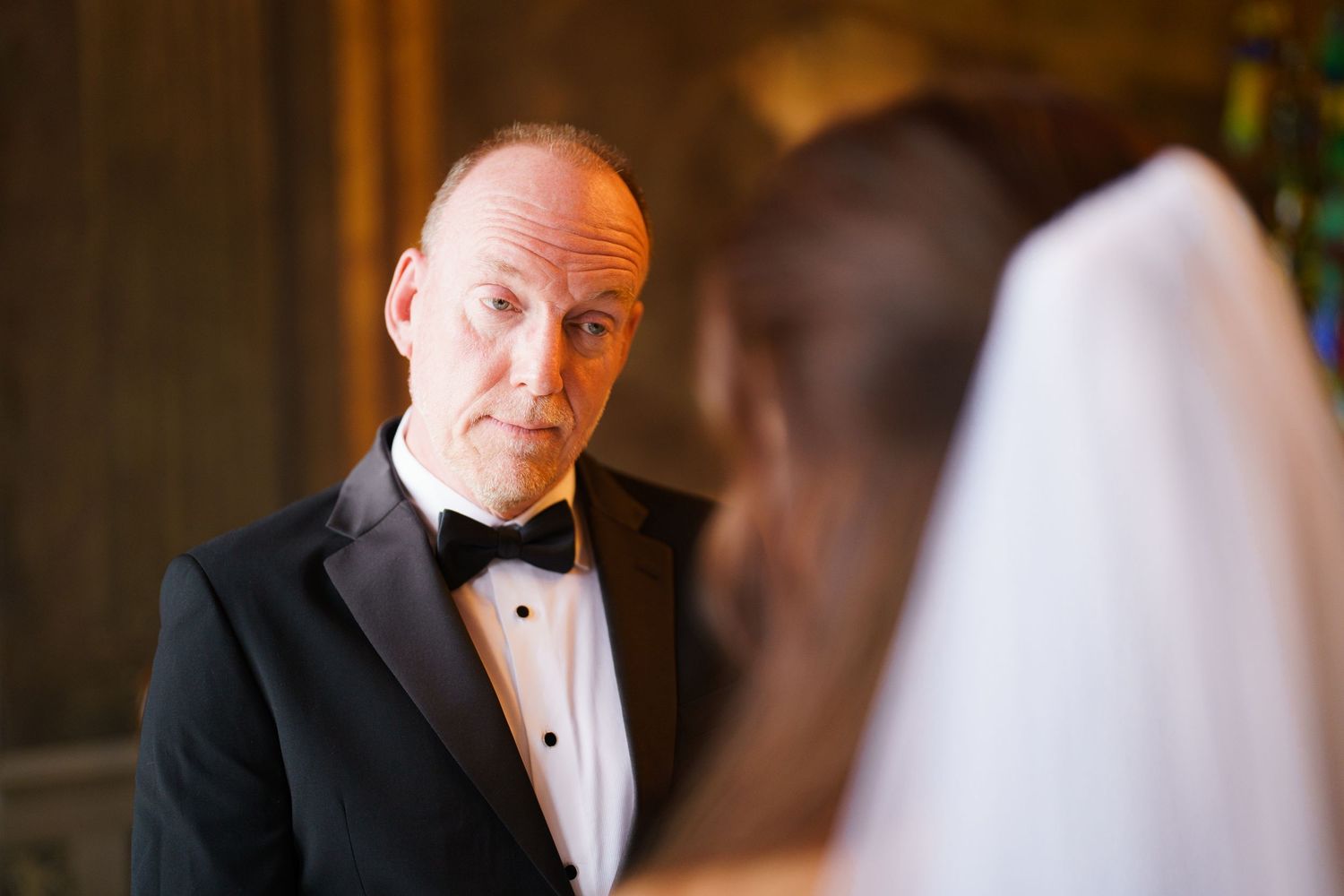 A person in a black tuxedo and bow tie stands in an elegant indoor setting during what appears to be a formal event.
