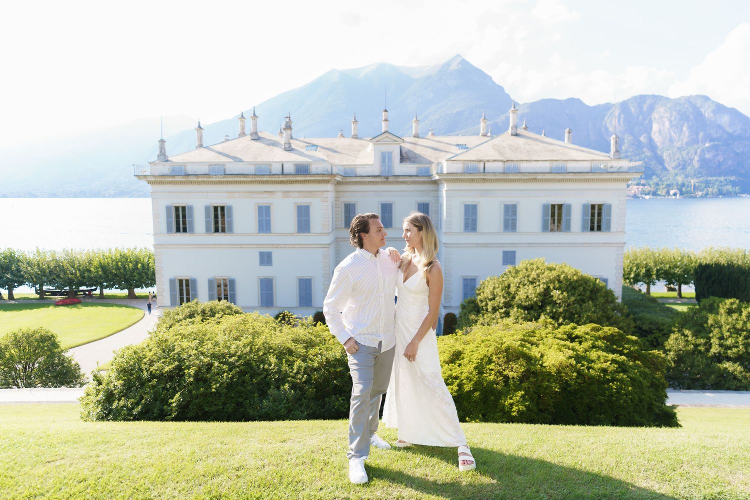 A couple in wedding attire stands on manicured lawn in front of elegant white villa with mountain views at Lake Como.