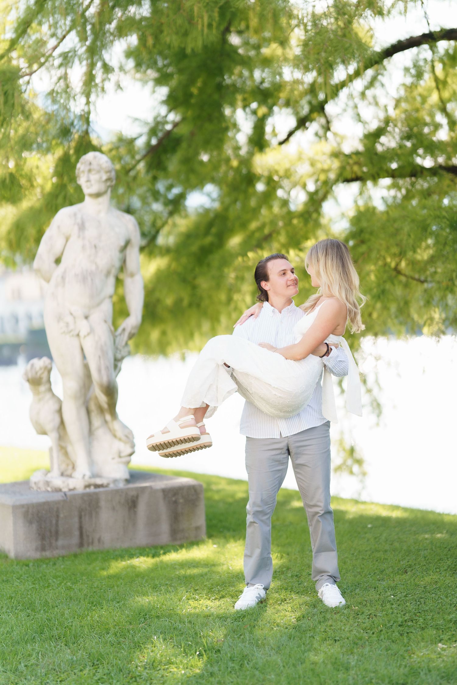 A couple shares a romantic moment dancing together on a grassy lawn near stone statues on a sunny afternoon.