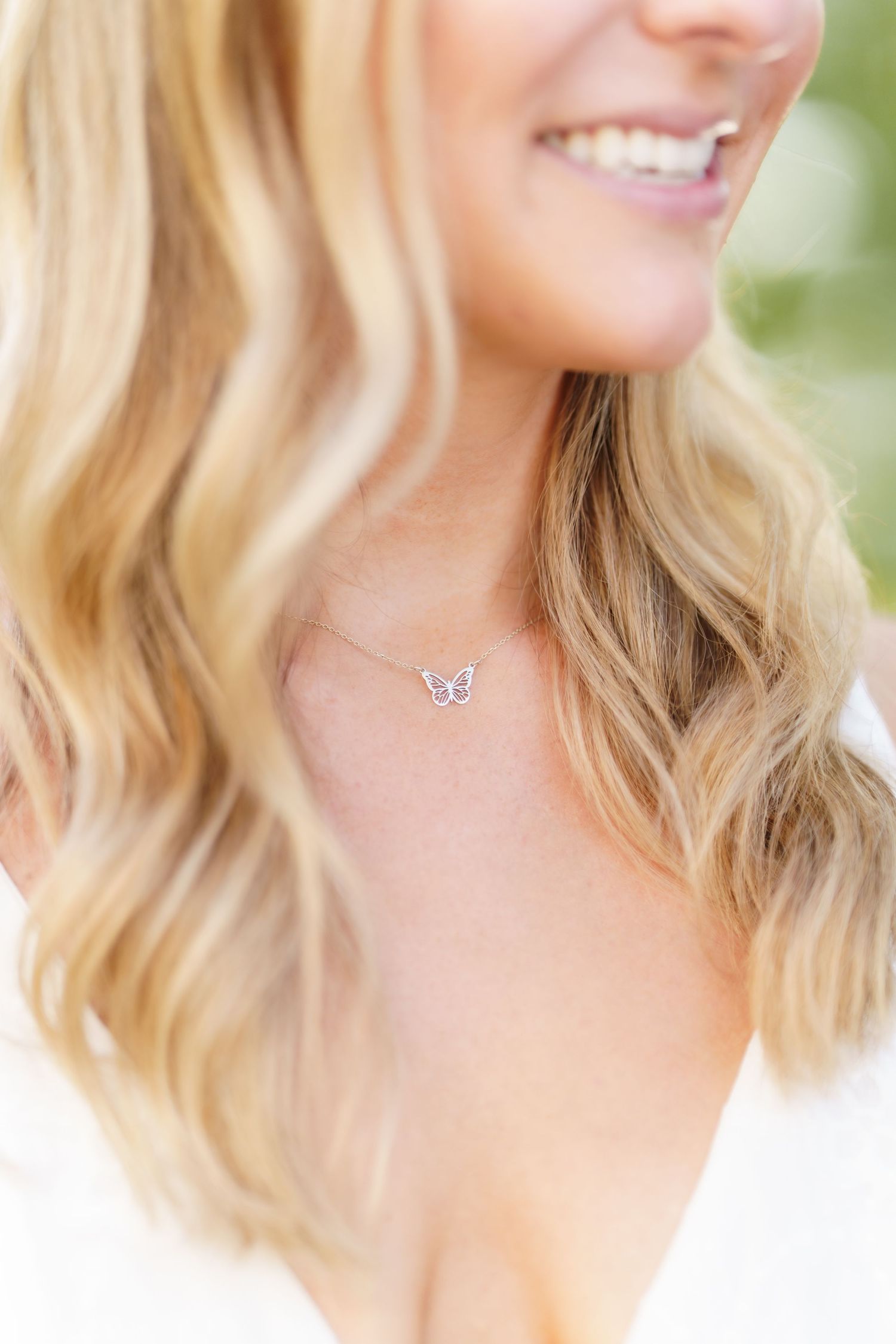 Close-up portrait of someone with wavy blonde hair and a bright smile wearing a delicate butterfly necklace.