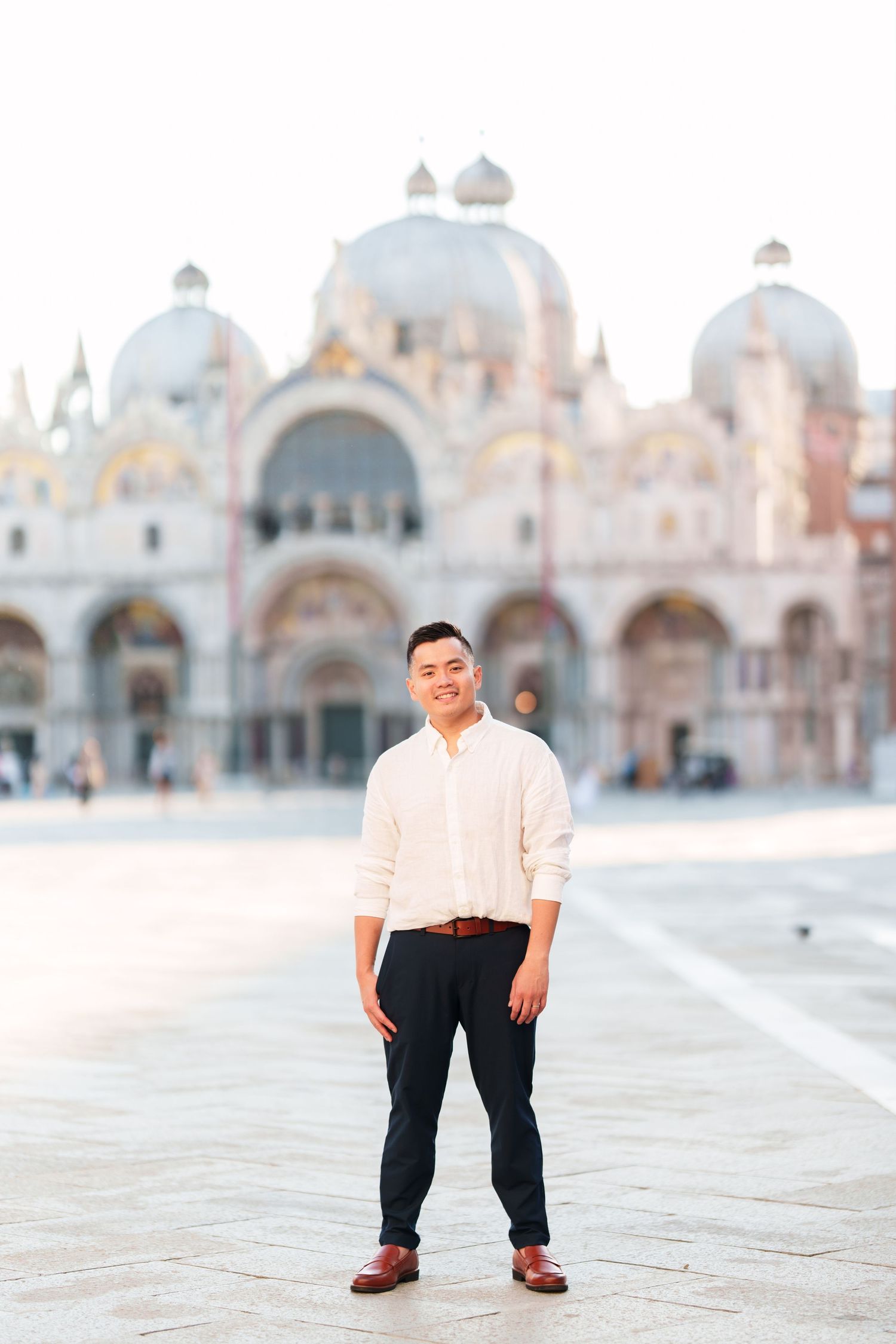 A smiling man in a white shirt and black pants stands in St. Mark's Square with the iconic Basilica in Venice, Italy.
