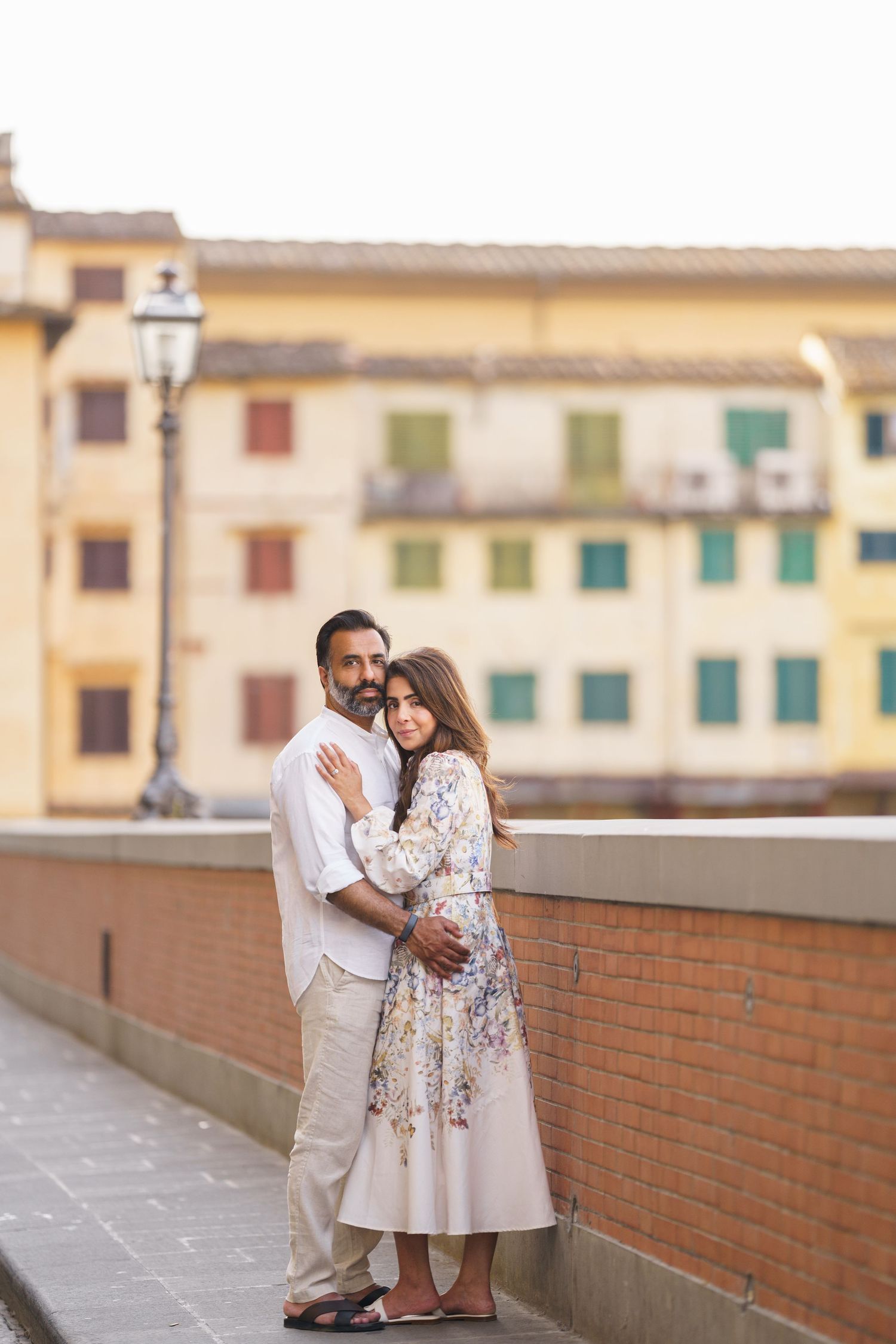 Couple embracing on a bridge in Florence, Italy, with colorful historic buildings in the background.