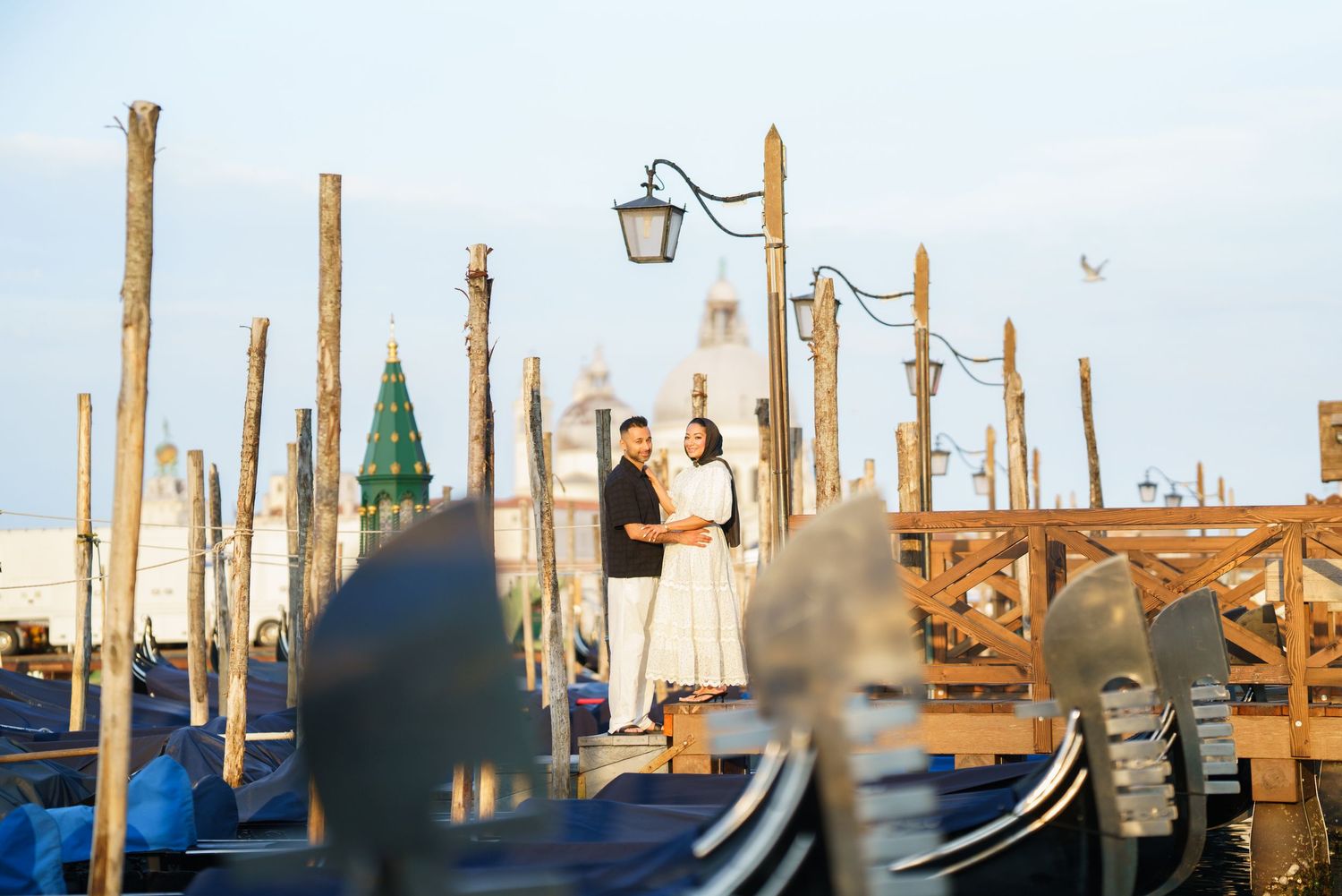 Couple embracing near gondolas and wooden posts at Venice waterfront with historic architecture backdrop.