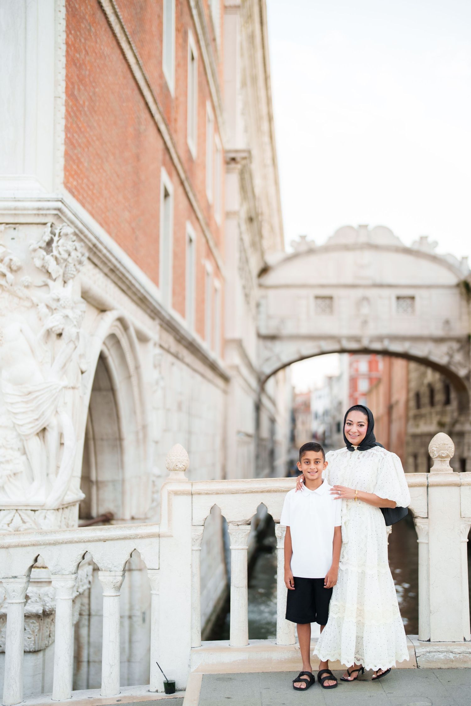 A woman in white hijab and dress poses with a young boy near the Bridge of Sighs in Venice, Italy.