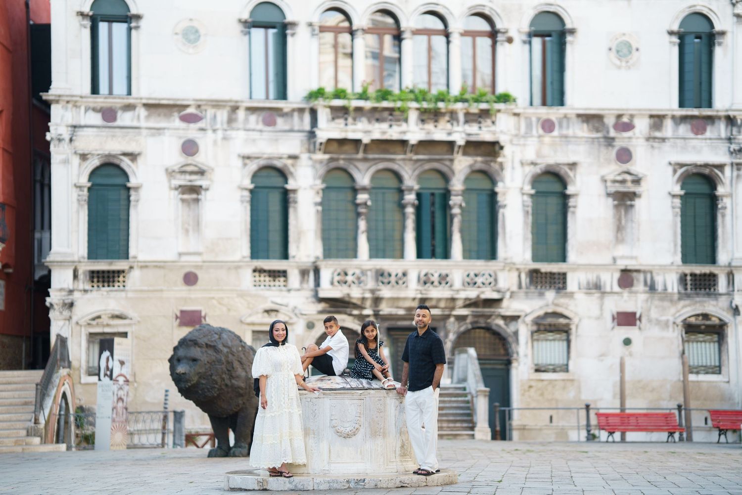 Family posing by a stone well in a Venice piazza with a historic ornate building and lion sculpture behind them.
