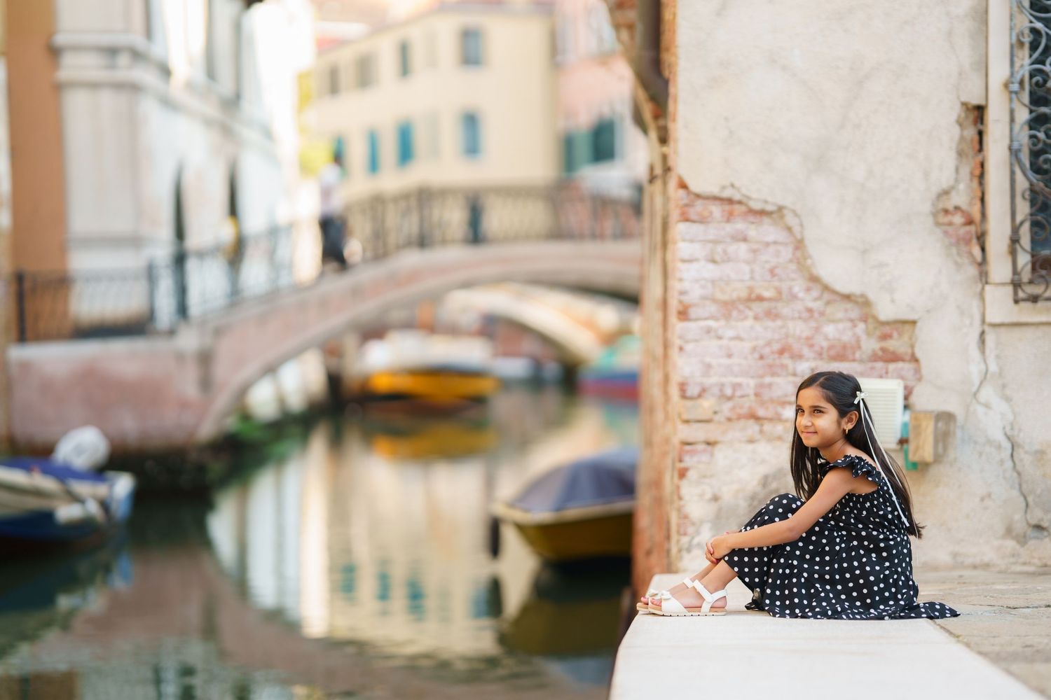 Young girl in polka dot dress sitting by a Venice canal with bridge and colorful boats in background.