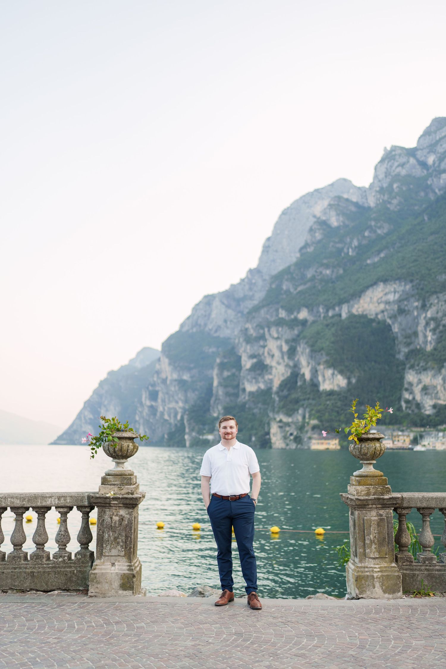 Man in white shirt and navy pants posing on lakeside terrace with dramatic mountain backdrop at Lake Garda.