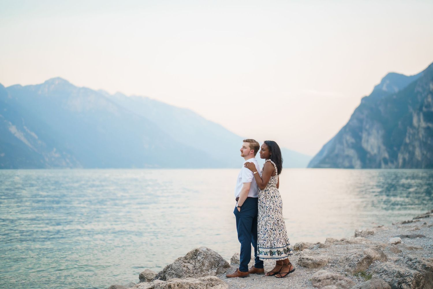 Couple standing on rocky shore of Lake Garda with misty mountains backdrop at dusk.