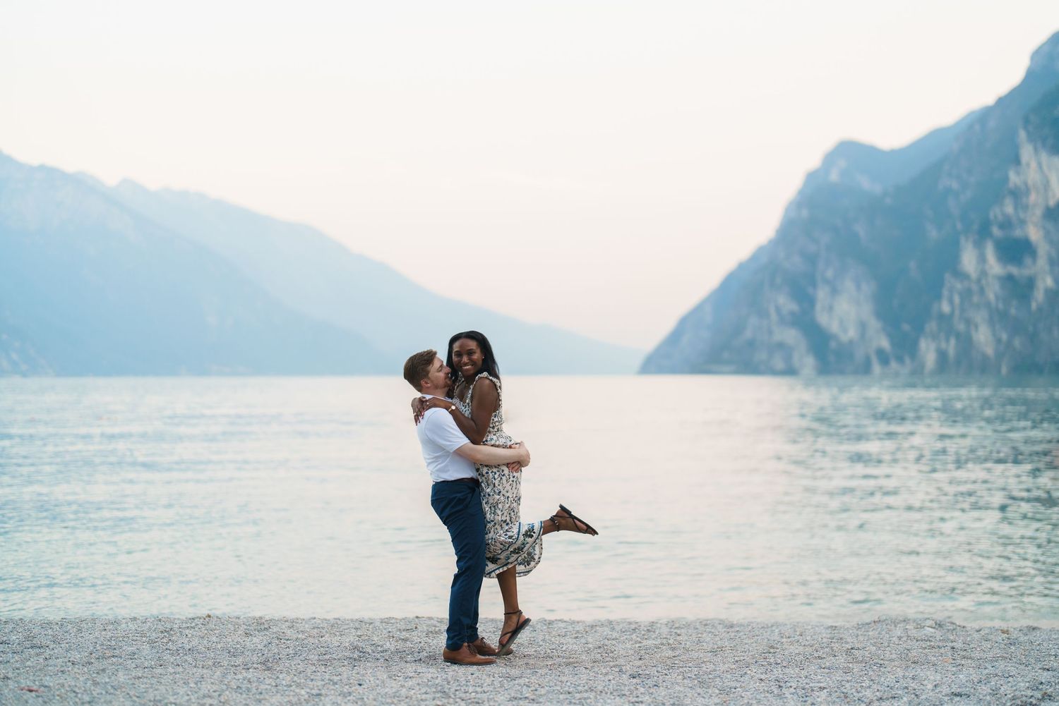Man lifting woman joyfully on Lake Garda beach with stunning mountain scenery behind.