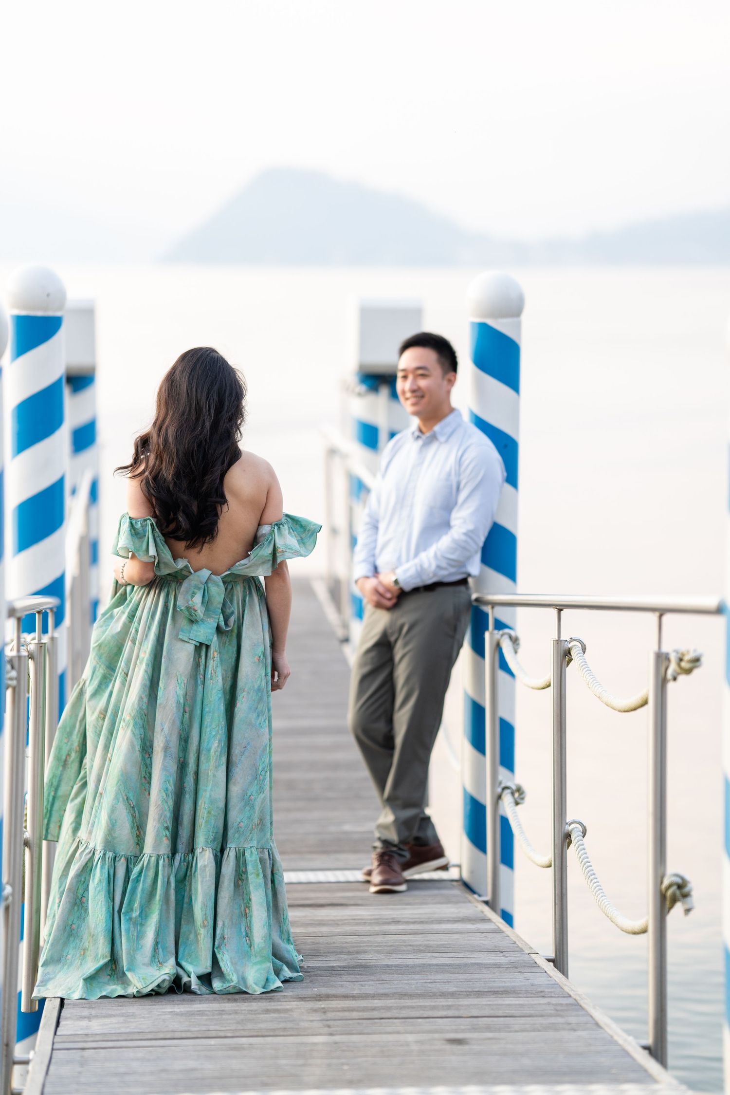 Couple on a seaside pier, woman in teal backless dress faces man in white shirt with ocean backdrop.