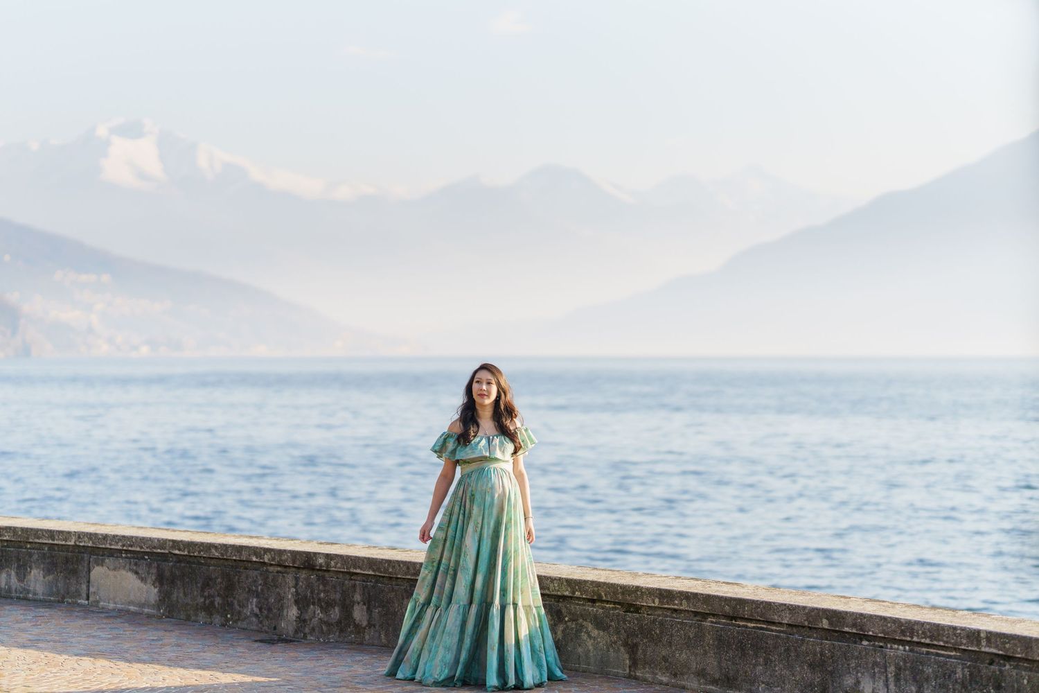 Woman in green gown poses on lakeside promenade with misty mountains and calm blue water in background.
