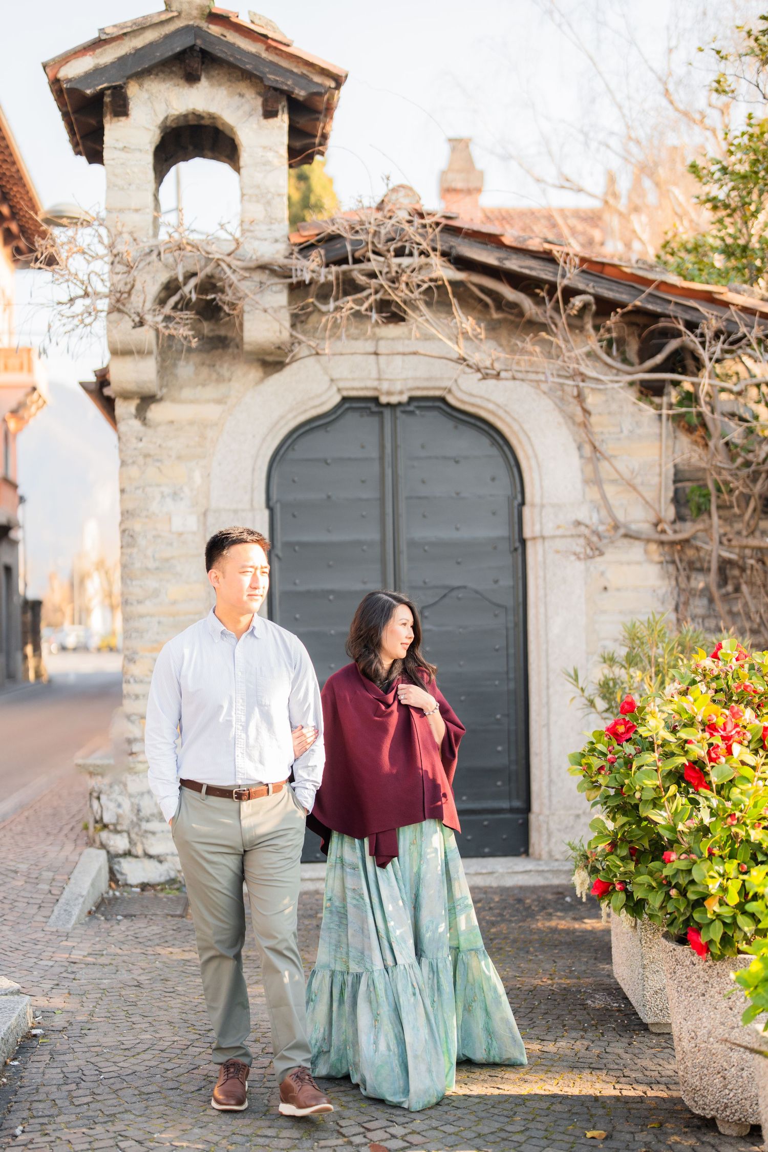 Couple posing in front of a stone archway door on a cobblestone street with red flowers nearby.