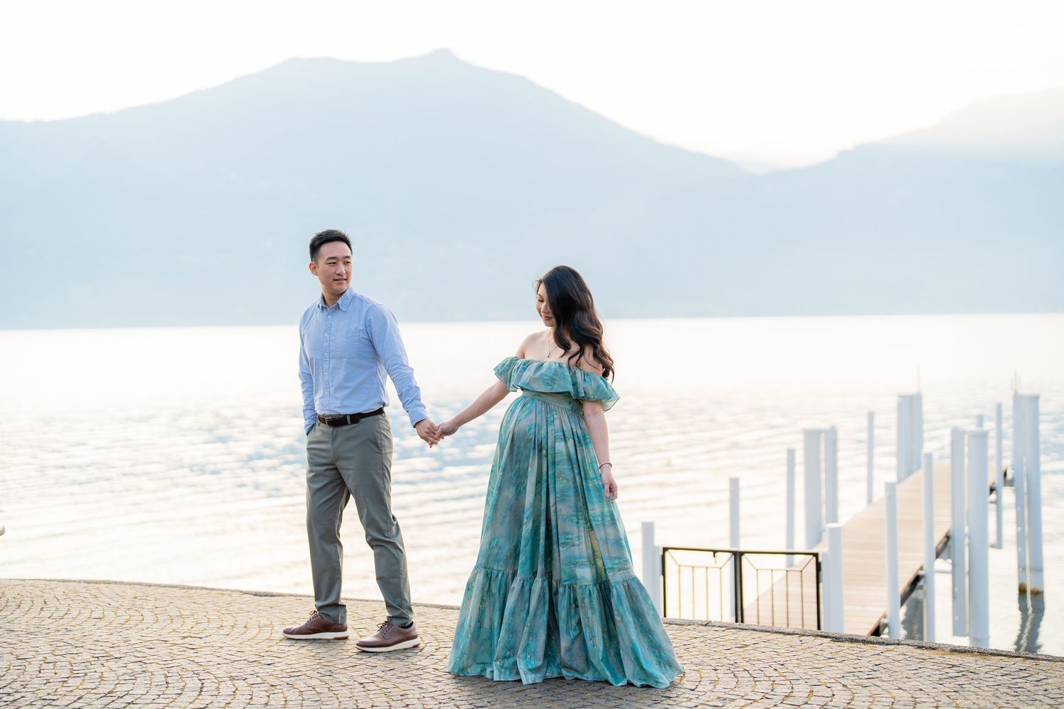 Couple holding hands on a lakeside promenade, woman in teal gown, mountains in background.