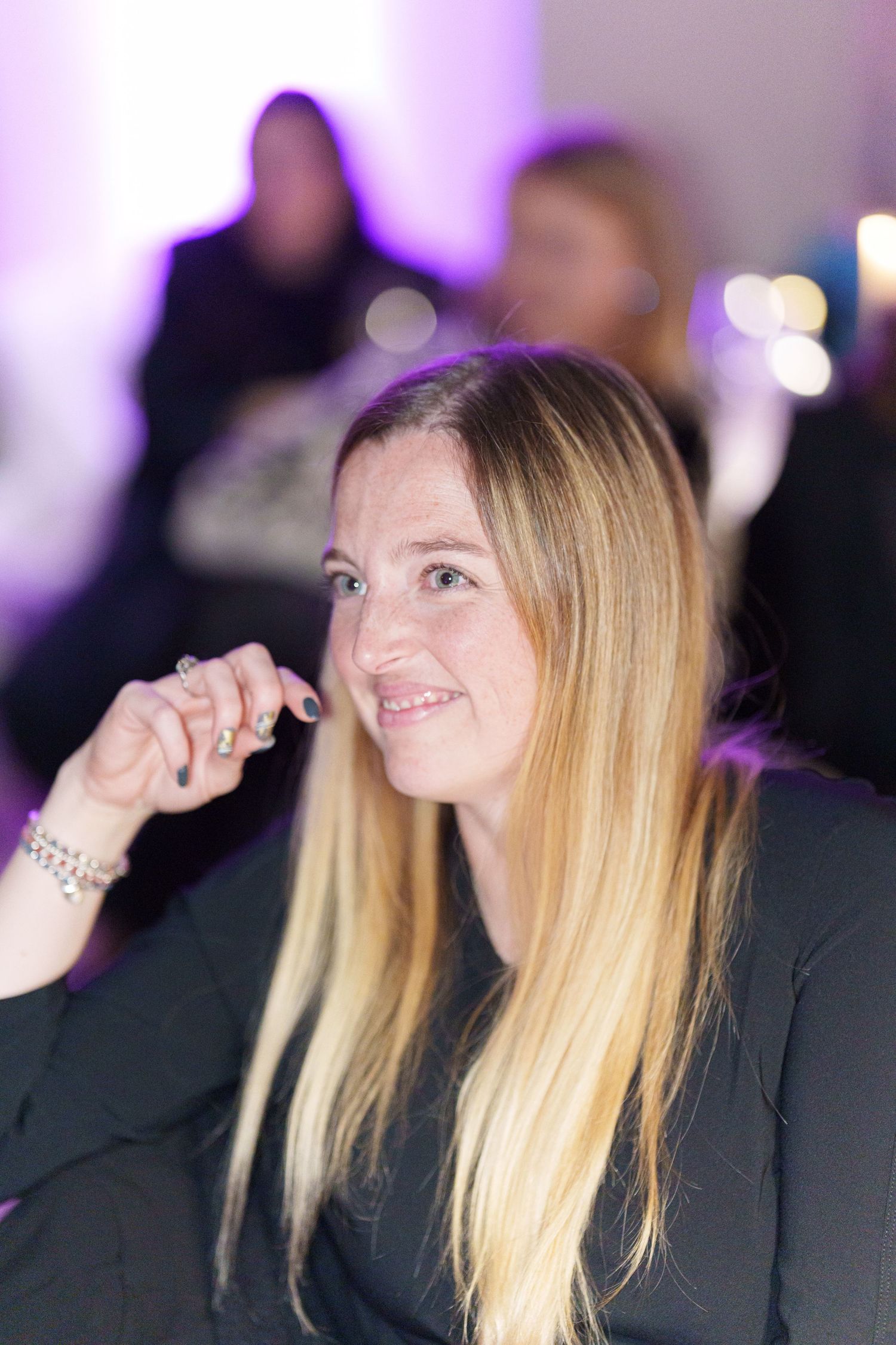 Smiling blonde woman at an event, hand raised to her chin, with purple ambient lighting in the background.